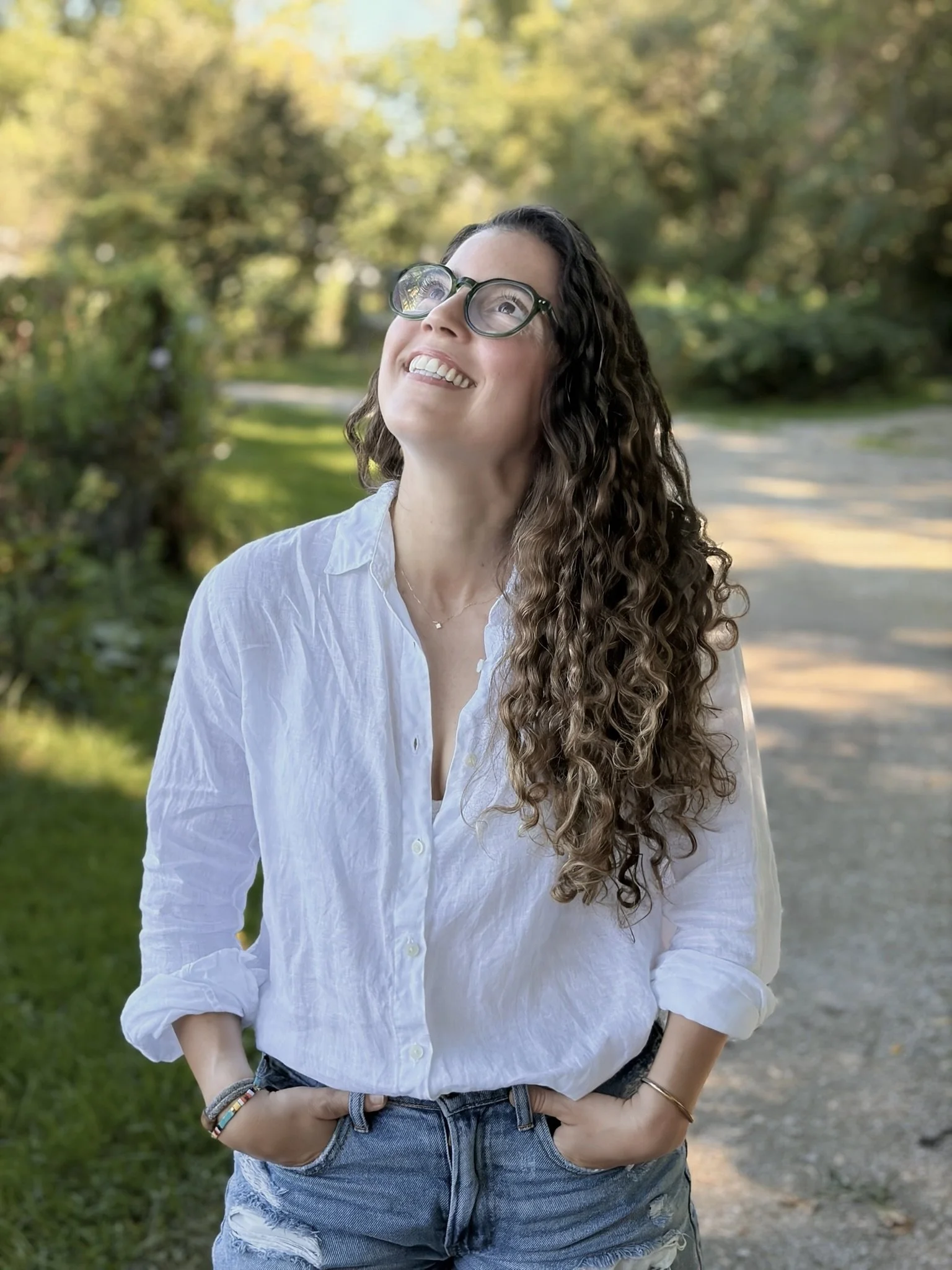 A young woman with curly brown hair and glasses smiling outdoors on a sunny day with a background of trees and a dirt path.
