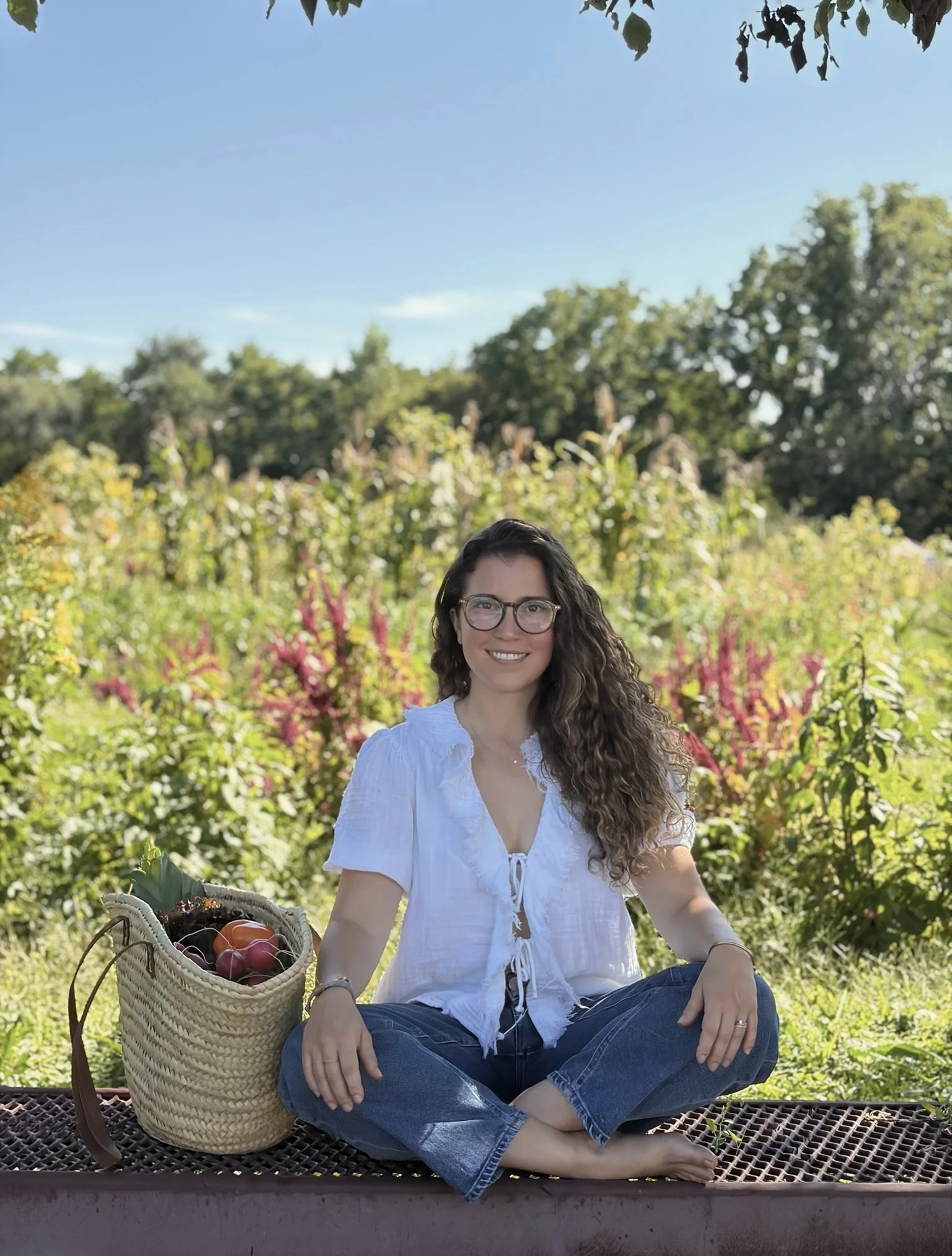 A woman with long curly hair, wearing glasses, a white blouse, and jeans, is sitting cross-legged on a basket bench outdoors in a garden with colorful flowers and trees, holding a basket of tomatoes and other vegetables.
