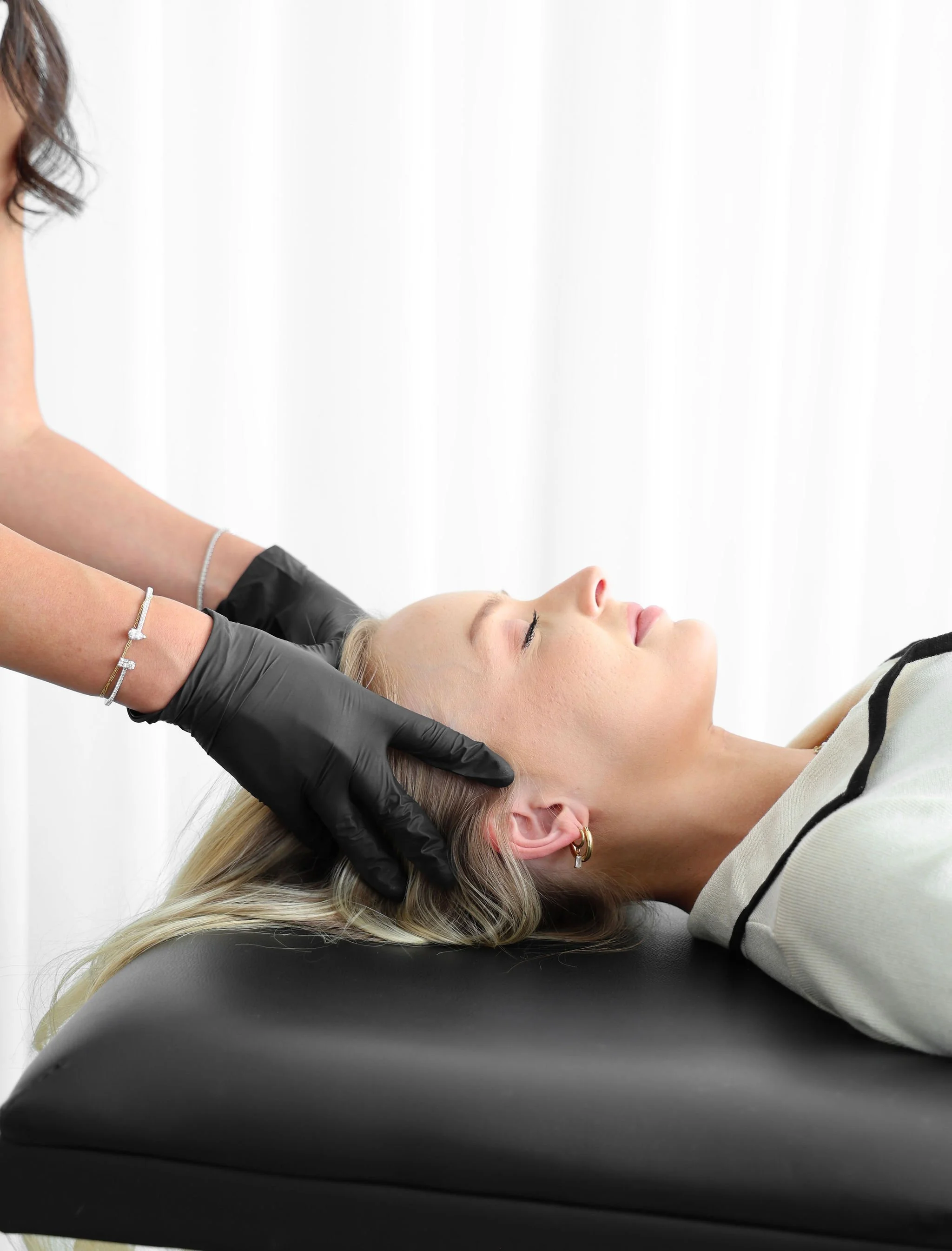 A woman lying back with her eyes closed on a black treatment chair, receiving a beauty or skincare procedure from a professional wearing black gloves.