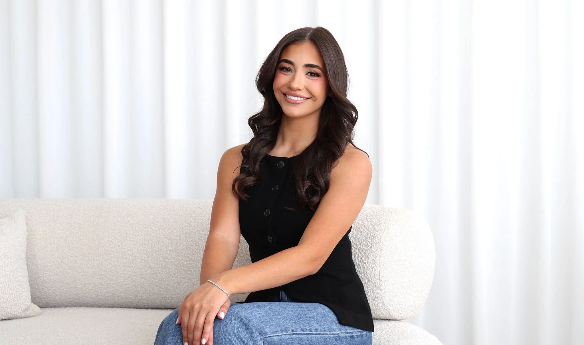 A young woman with long dark hair and a black sleeveless top sitting on a light-colored sofa in front of white curtains, smiling at the camera.