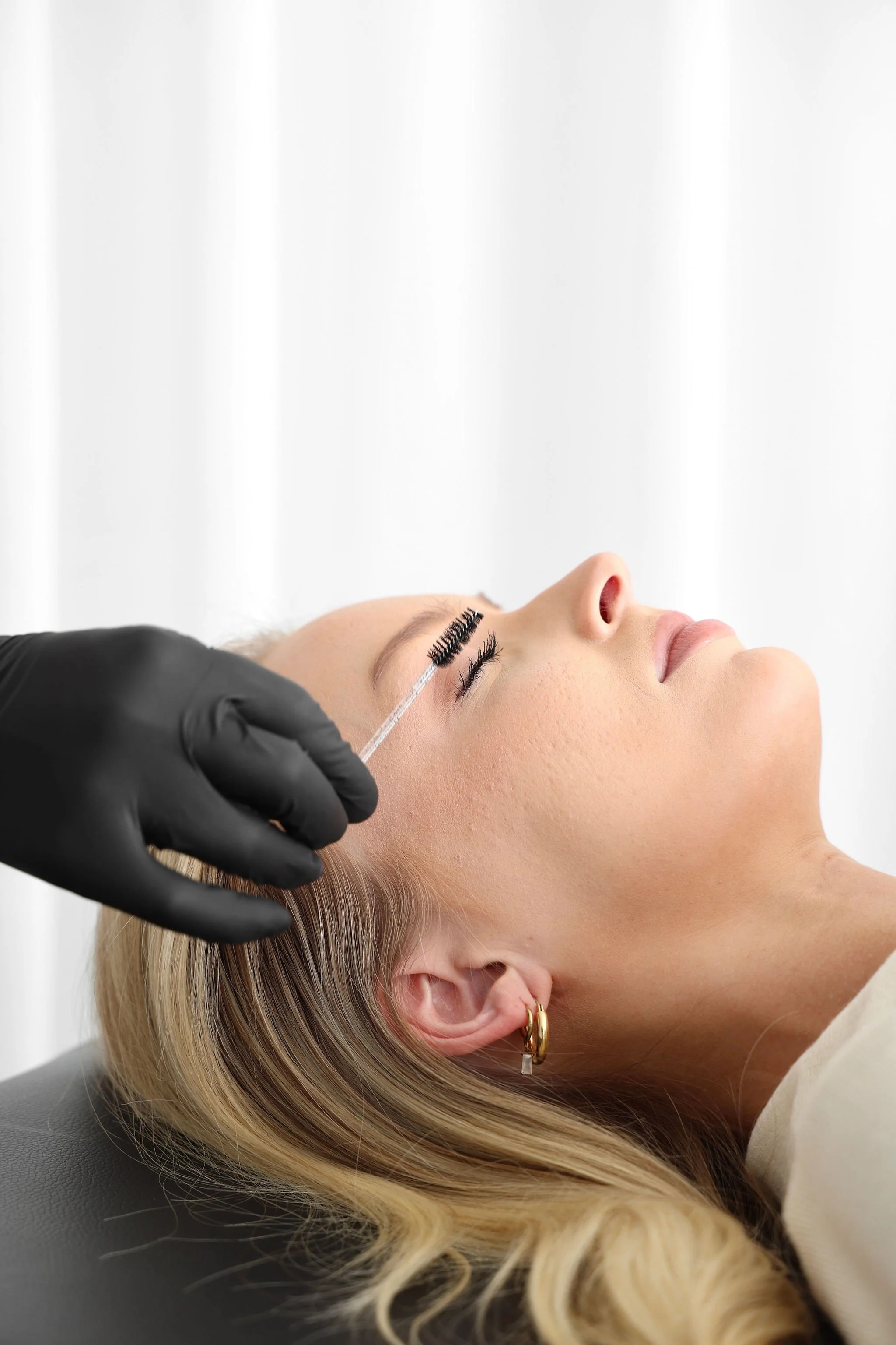 Woman lying down with eyes closed, receiving eyelash extension application, eyelash technician in black glove using a tool on her eyelashes.