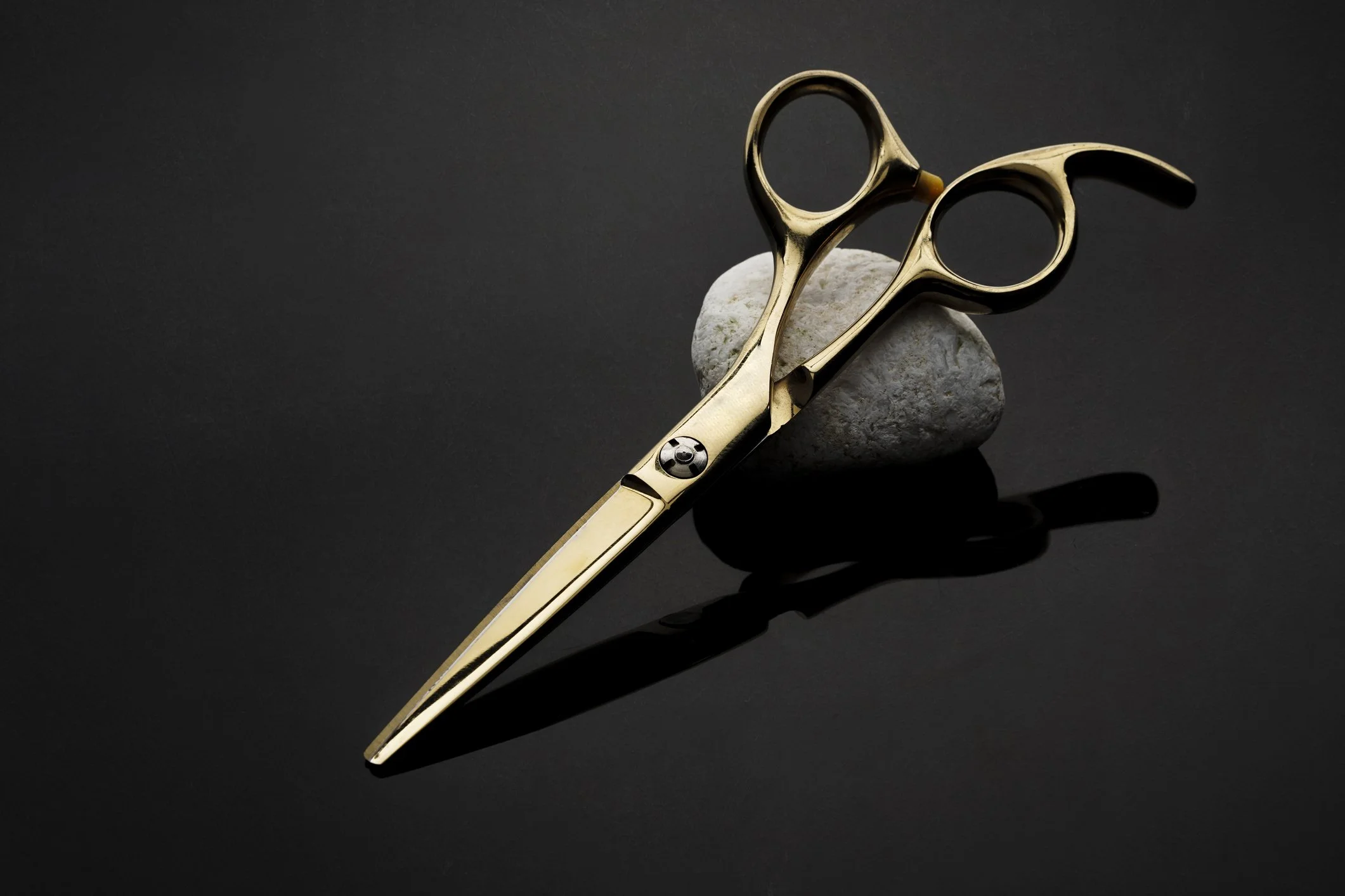 Gold barber scissors resting on dark stones against a black background.