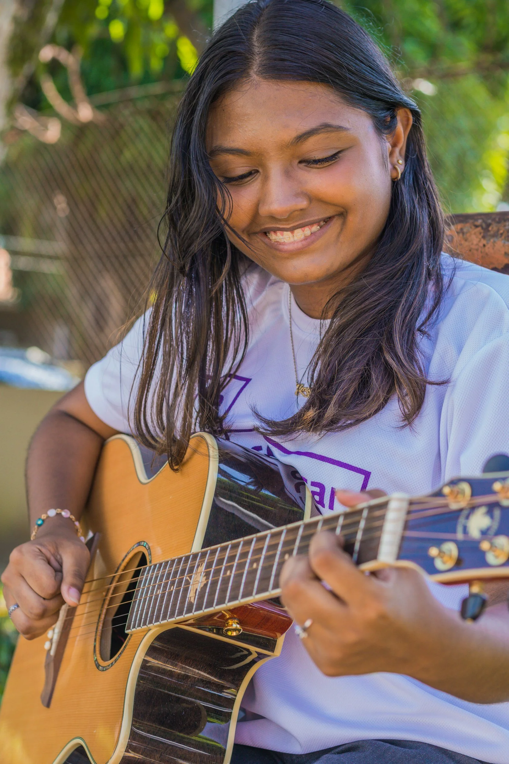 A young woman with long dark hair, wearing earrings and a white T-shirt, playing an acoustic guitar outdoors, smiling and looking down, with green foliage in the background.