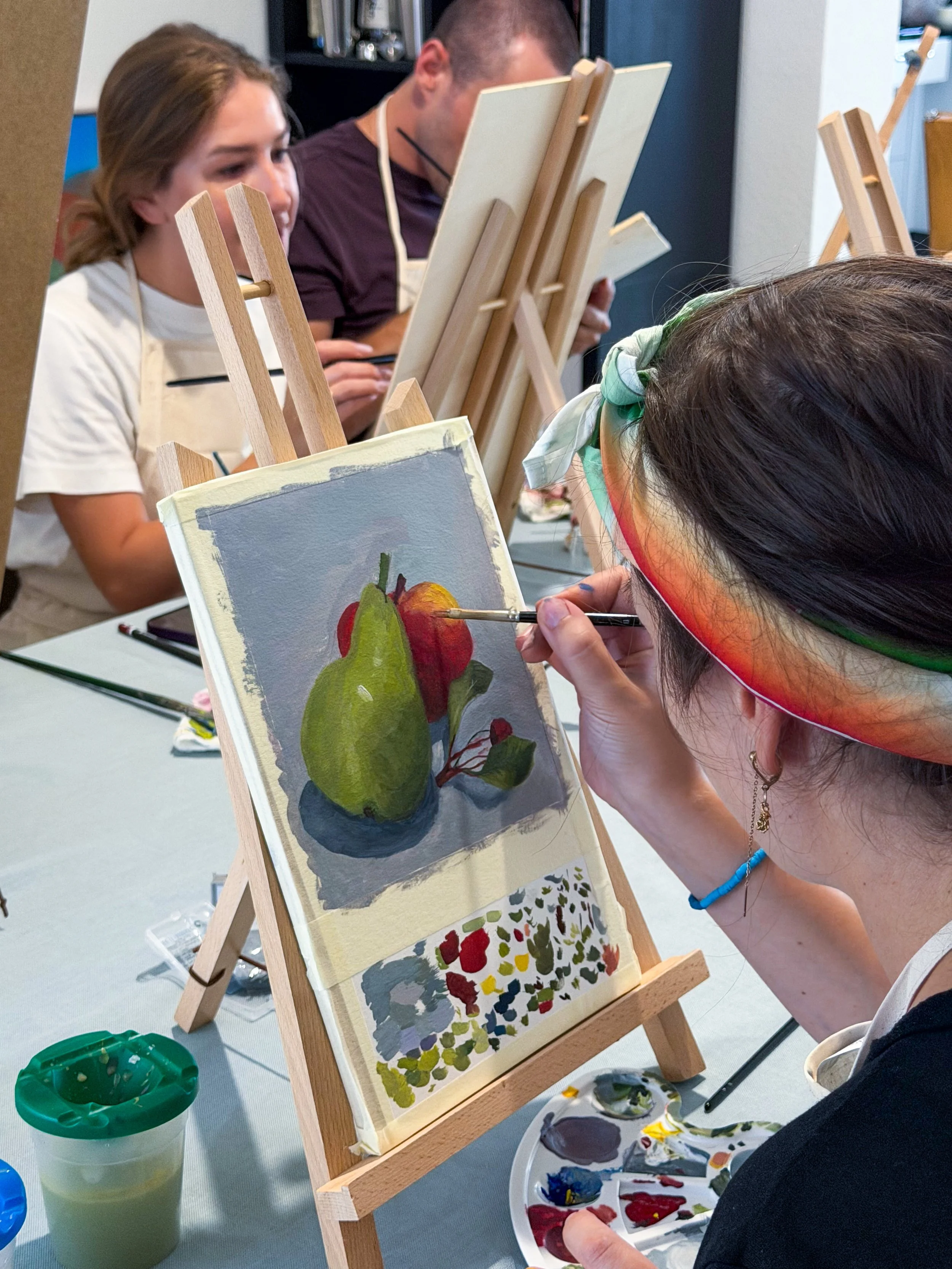 People painting still life art on canvas at an art class, with easels and paint palettes on the table.