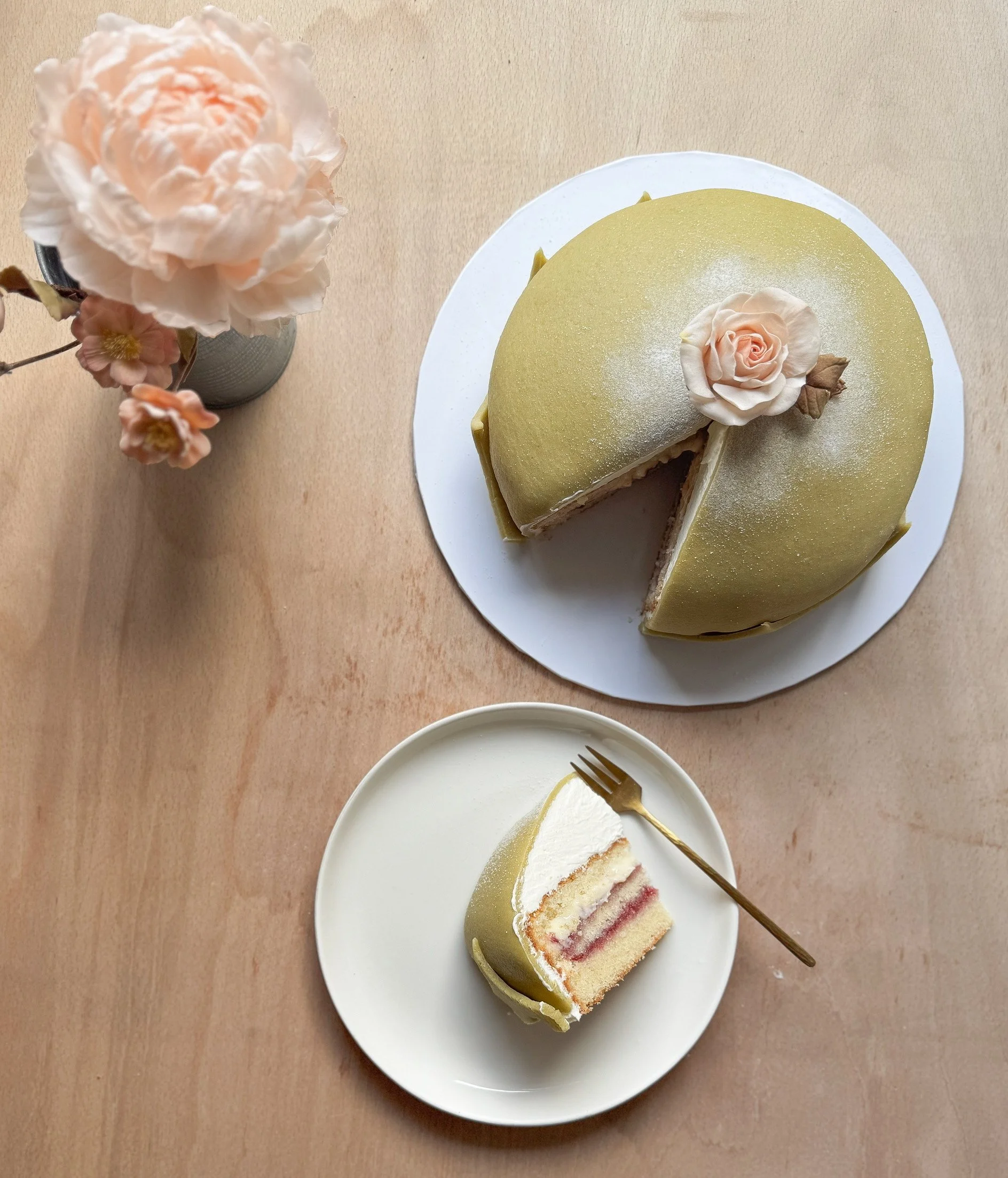 A three-layer sponge cake with white frosting and pink filling, decorated with a pink and white flower on top, on a white plate and a gold fork, with a second slice cut out and placed on a separate white plate, decorated with a pink flower and a smaller flower in a vase nearby.