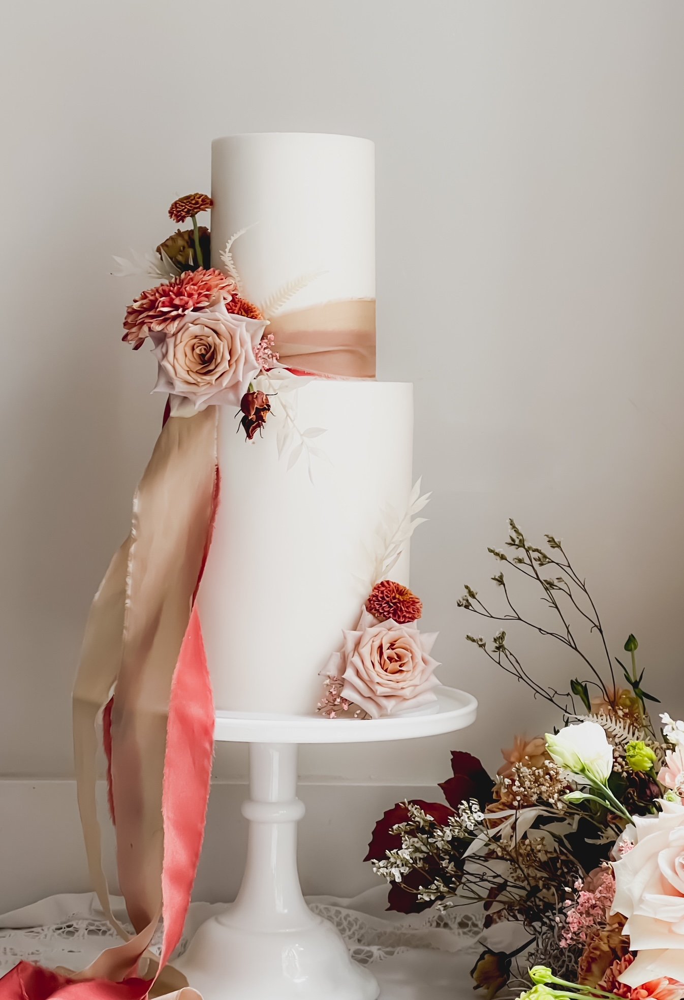 A white, two-tier cake decorated with pink and peach roses, ribbons, and floral accents, displayed on a white cake stand next to a bouquet of assorted flowers.