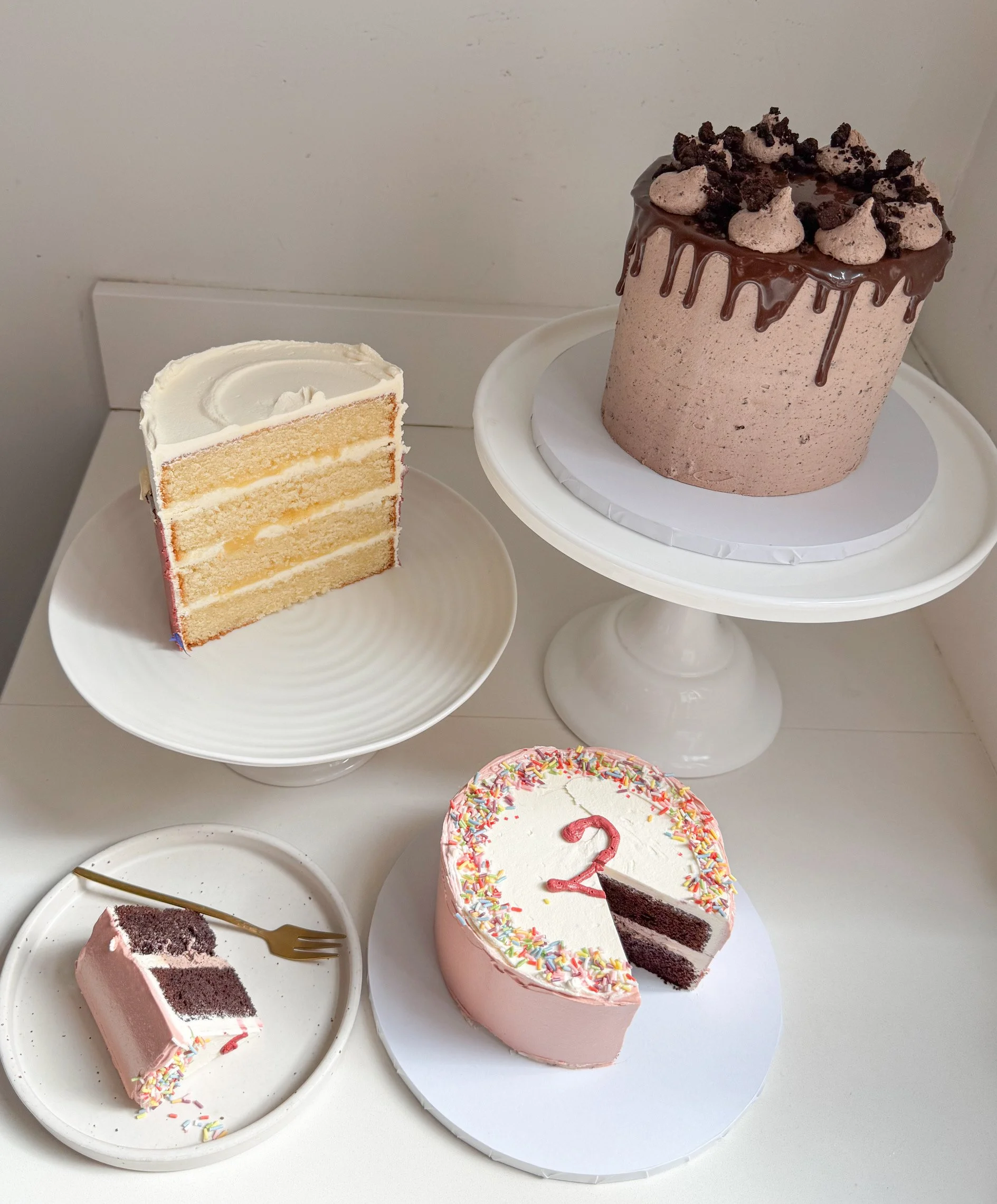A large chocolate birthday cake with pink icing and rainbow sprinkles, decorated with buttercream and cookie crumbles on top, with a slice removed. Next to it is a slice of vanilla cake with white frosting on a white plate.