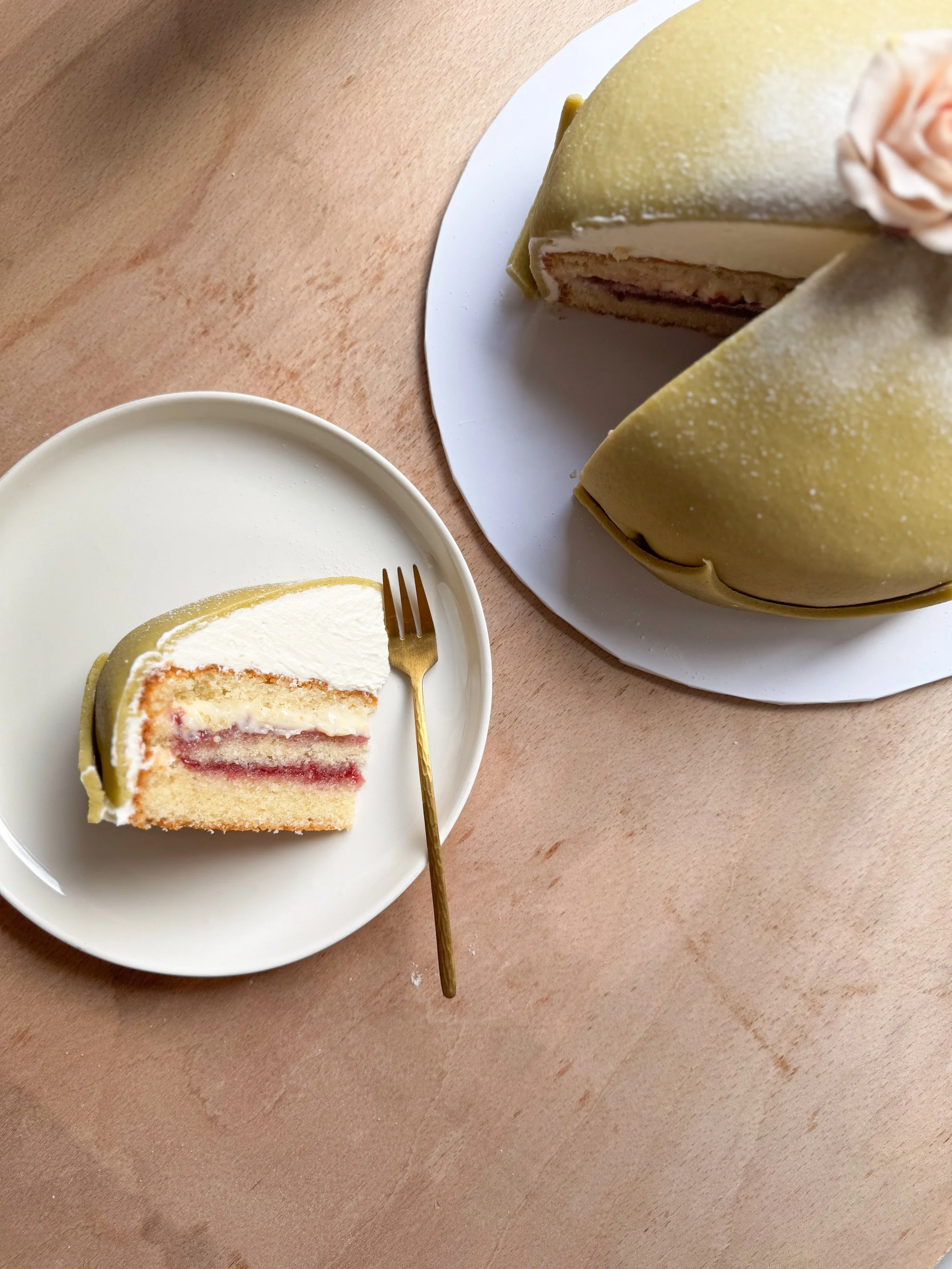 A slice of layered cake with cream and fruit filling on a white plate, with a cake whole with yellow glaze and a pink flower decoration on top, on a wooden surface, and a bowl of white icing or cream nearby.