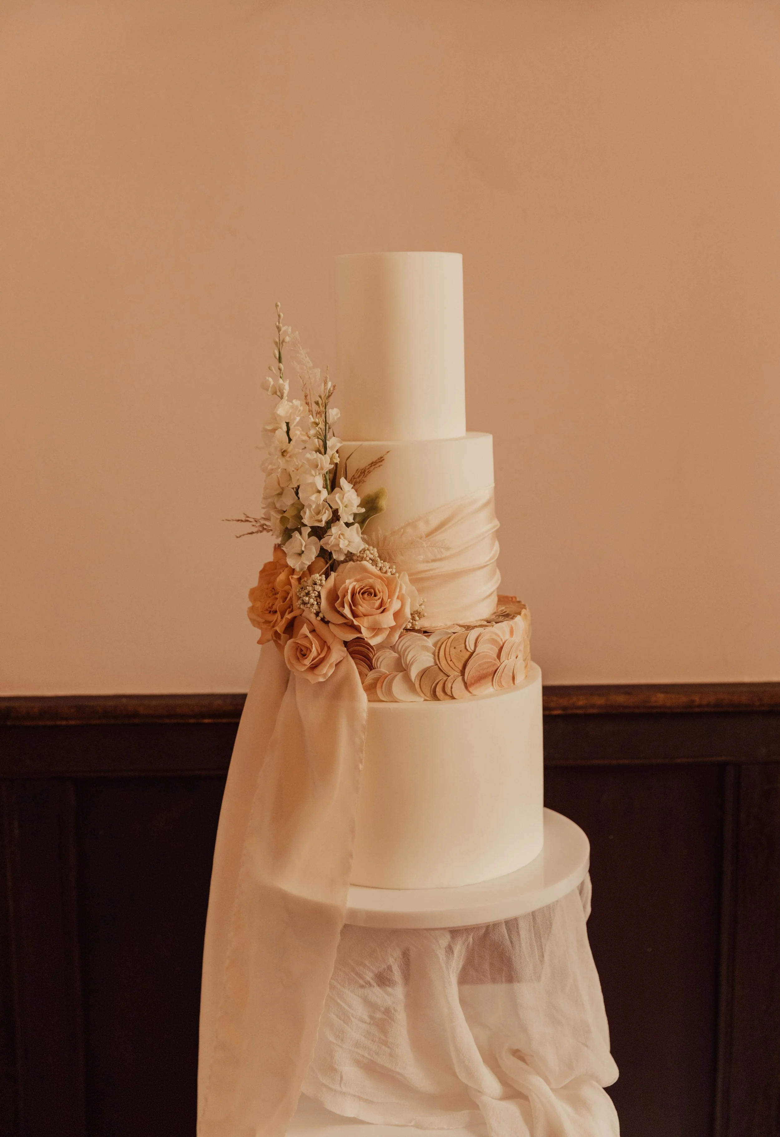 A white wedding cake with four tiers decorated with fresh flowers and fabric, set against a peach-colored background.