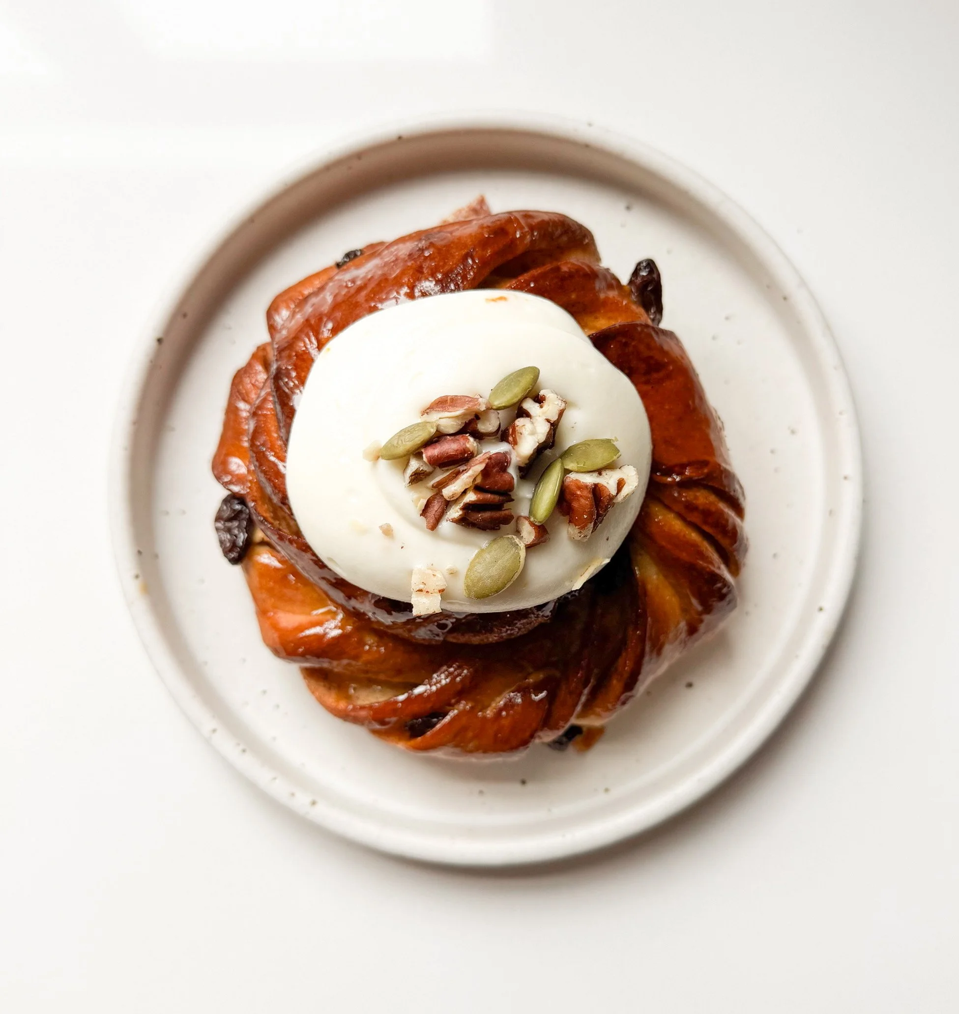 Close-up of a dessert with a white round topping, pumpkin seeds, chopped nuts, and dried fruits on a white plate.
