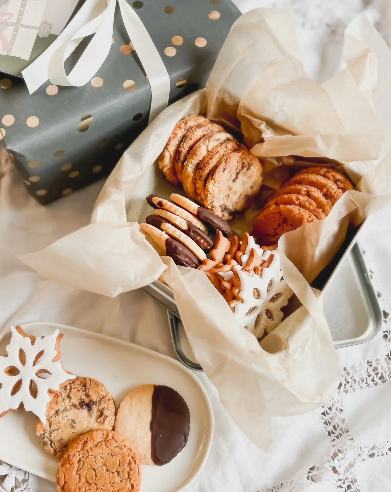 A gift box with white ribbon sits on a table near a tin container with assorted Christmas cookies, some dipped in chocolate, on a lace tablecloth.