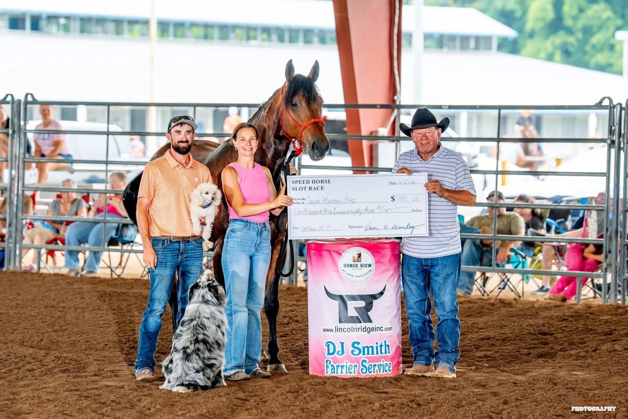 A group of people at a horse racing event holding a large check with a racing horse and two dogs present. The scene is set in an indoor arena with spectators in the background.