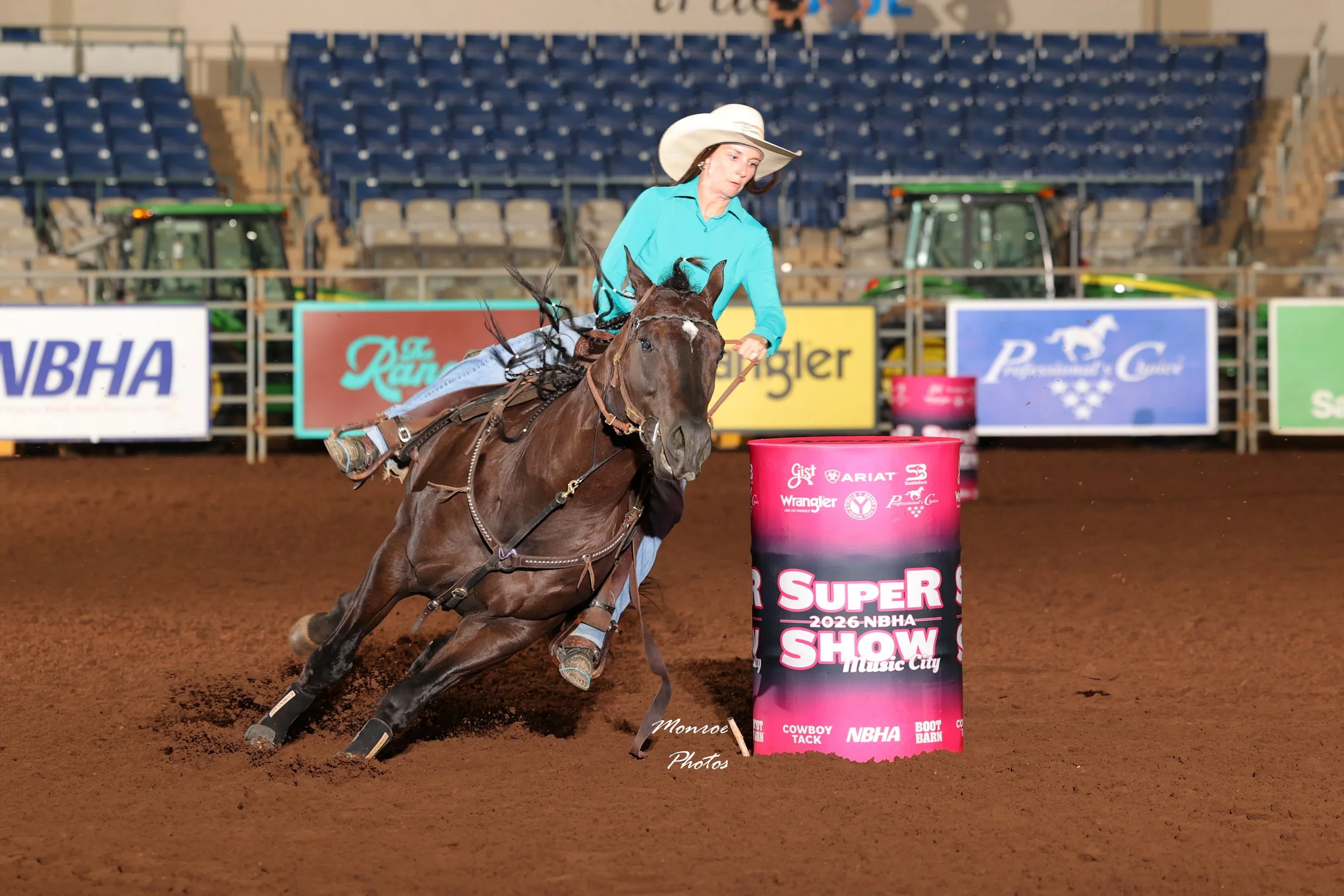 A female barrel racer wearing a wide-brimmed hat and turquoise shirt navigates a barrel in an indoor arena during a rodeo event, with empty stands and various sponsor banners in the background.