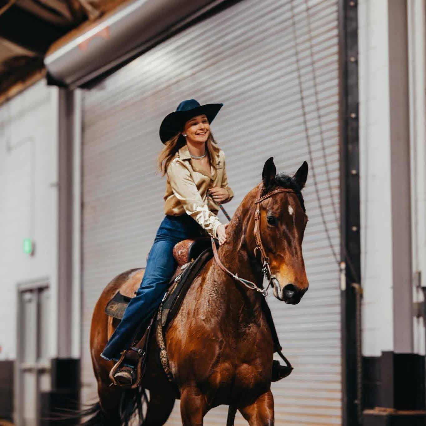 A woman riding a brown horse inside a indoor riding arena, smiling and wearing a black wide-brimmed hat, a beige shiny shirt, and blue jeans.