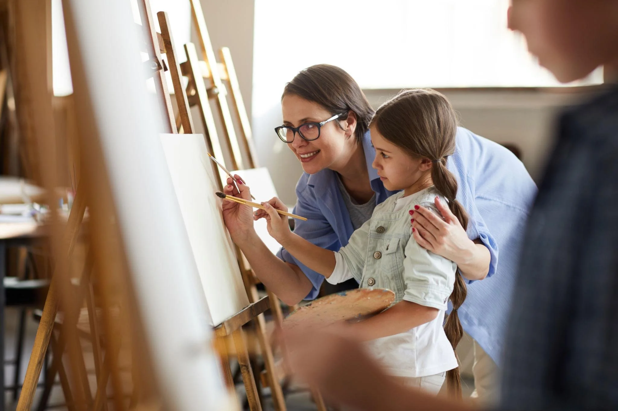A woman and a young girl are painting on a blank canvas during an art class in a bright room, with easels and other art supplies around them.