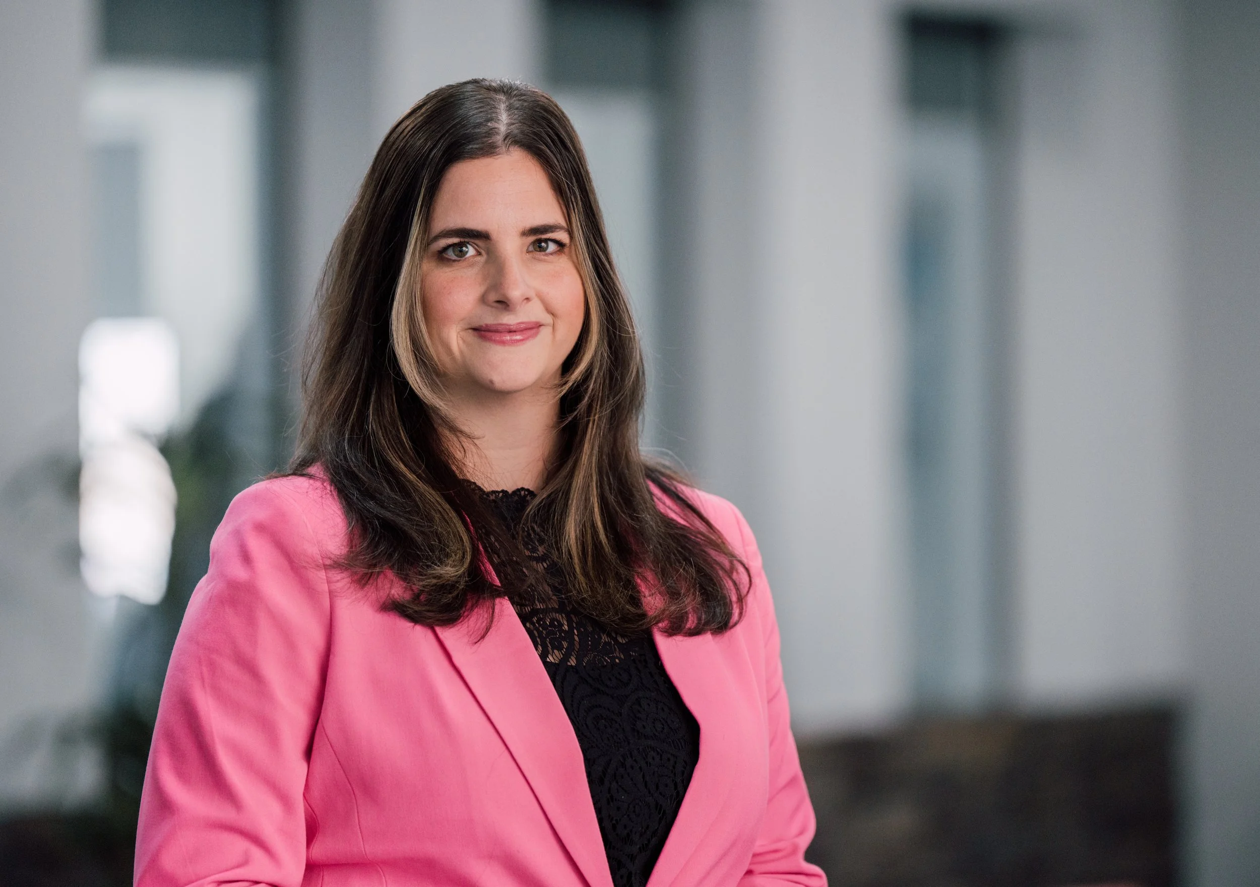 A woman with long brown hair, wearing a pink blazer over a black lace top, smiling softly, standing in front of a blurred modern building background.
