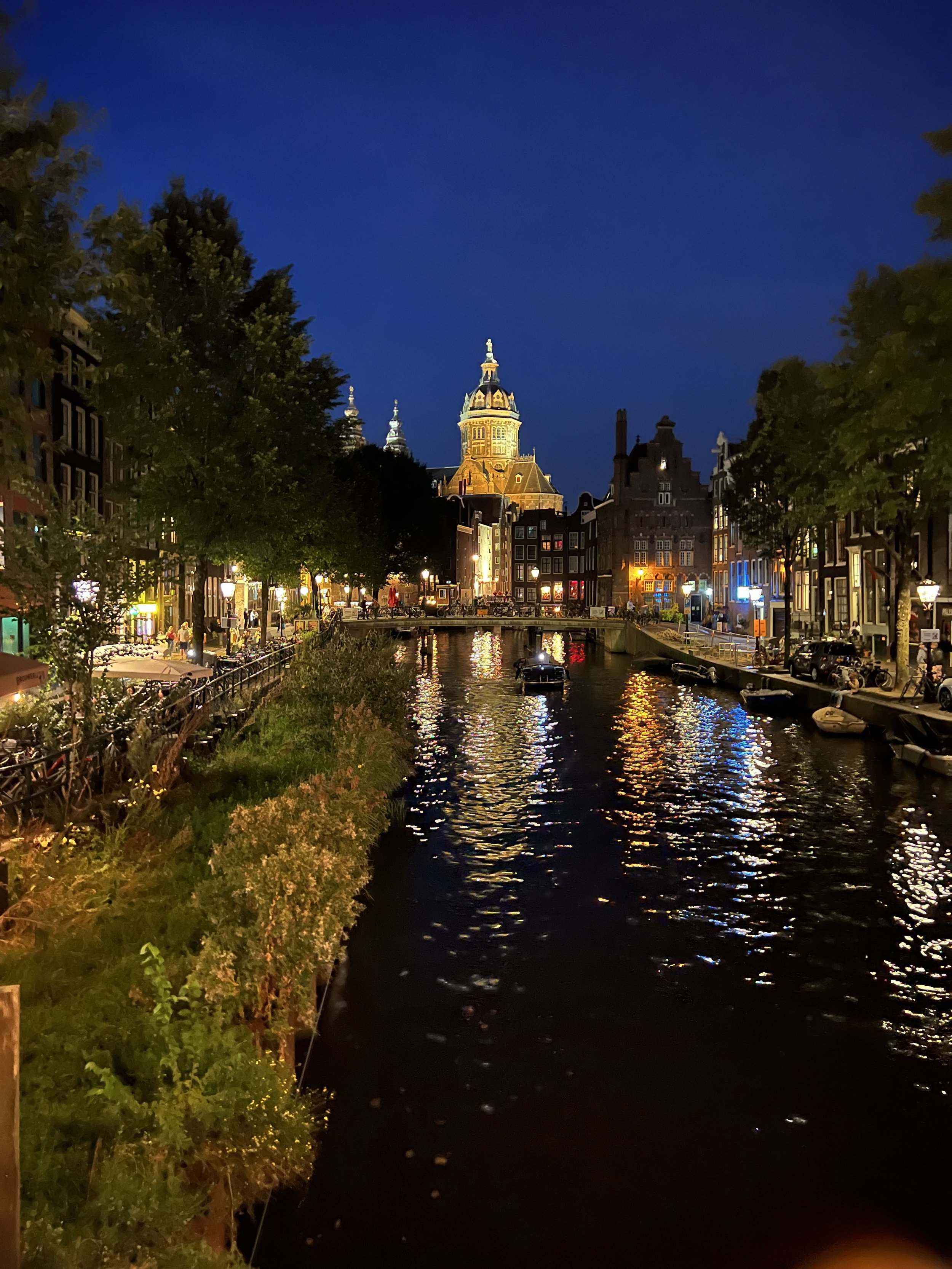 Night view of a canal with boat reflections, illuminated buildings, a tall church or historic tower, trees along the water, and a clear dark blue sky.