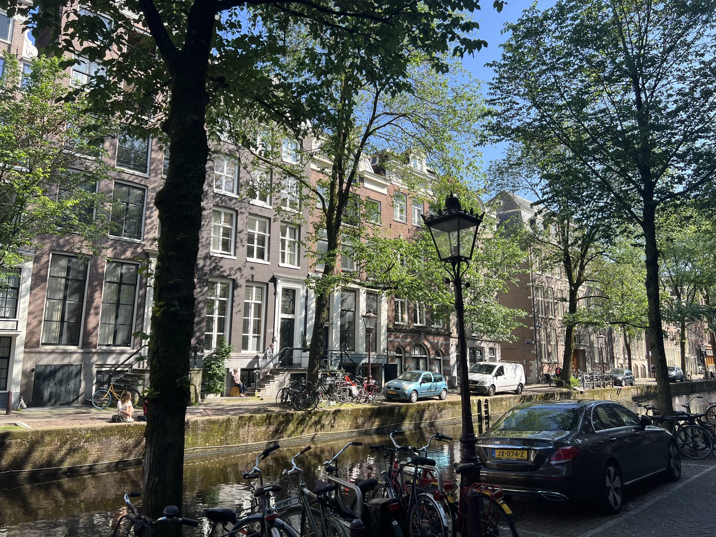 City street scene with tall brick buildings, green trees, parked bicycles, cars, a canal, and a person sitting on the sidewalk.