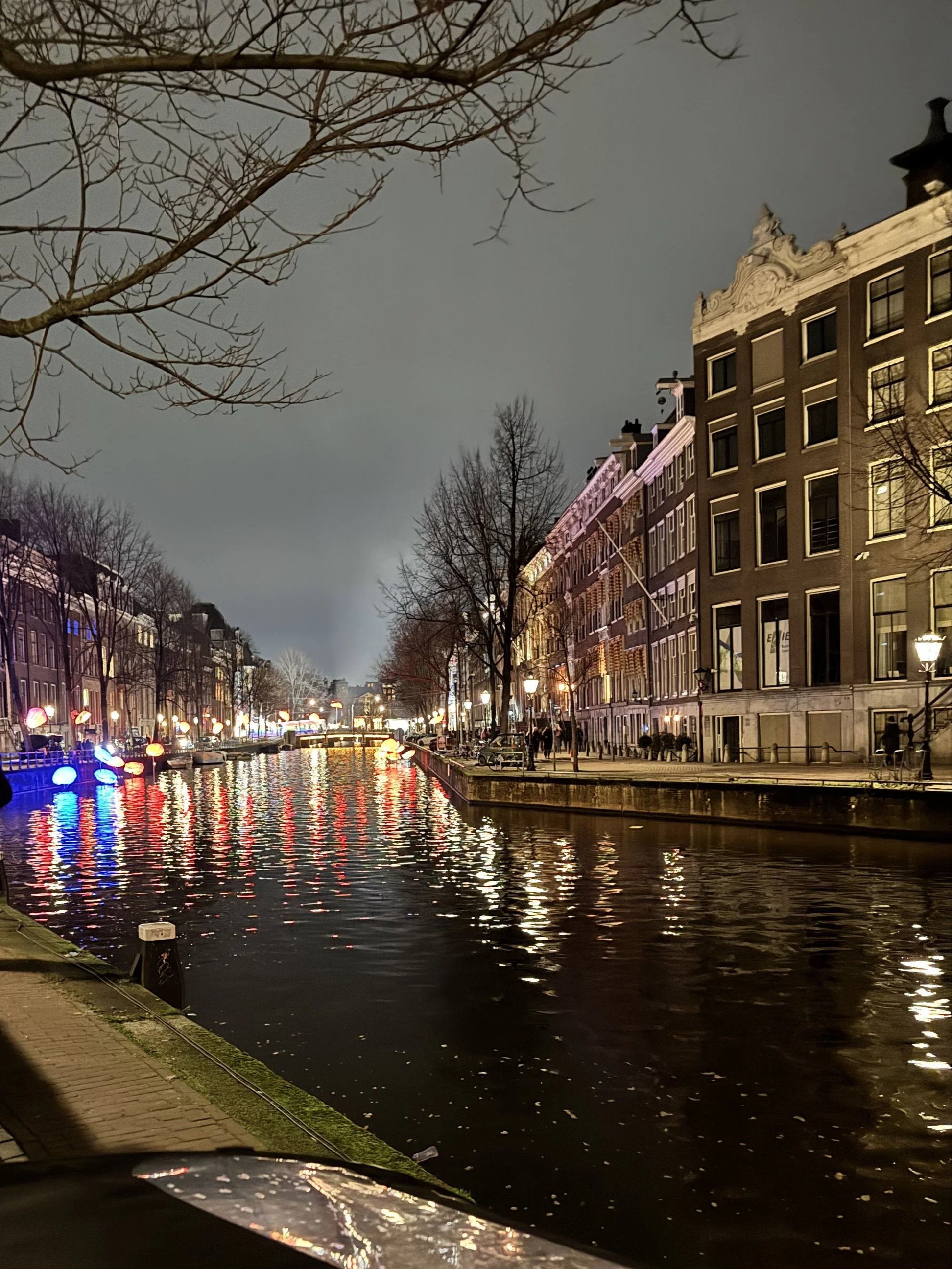 Nighttime scene of a canal with illuminated boats reflecting on the water, flanked by historic buildings and leafless trees, with a gray sky in the background.