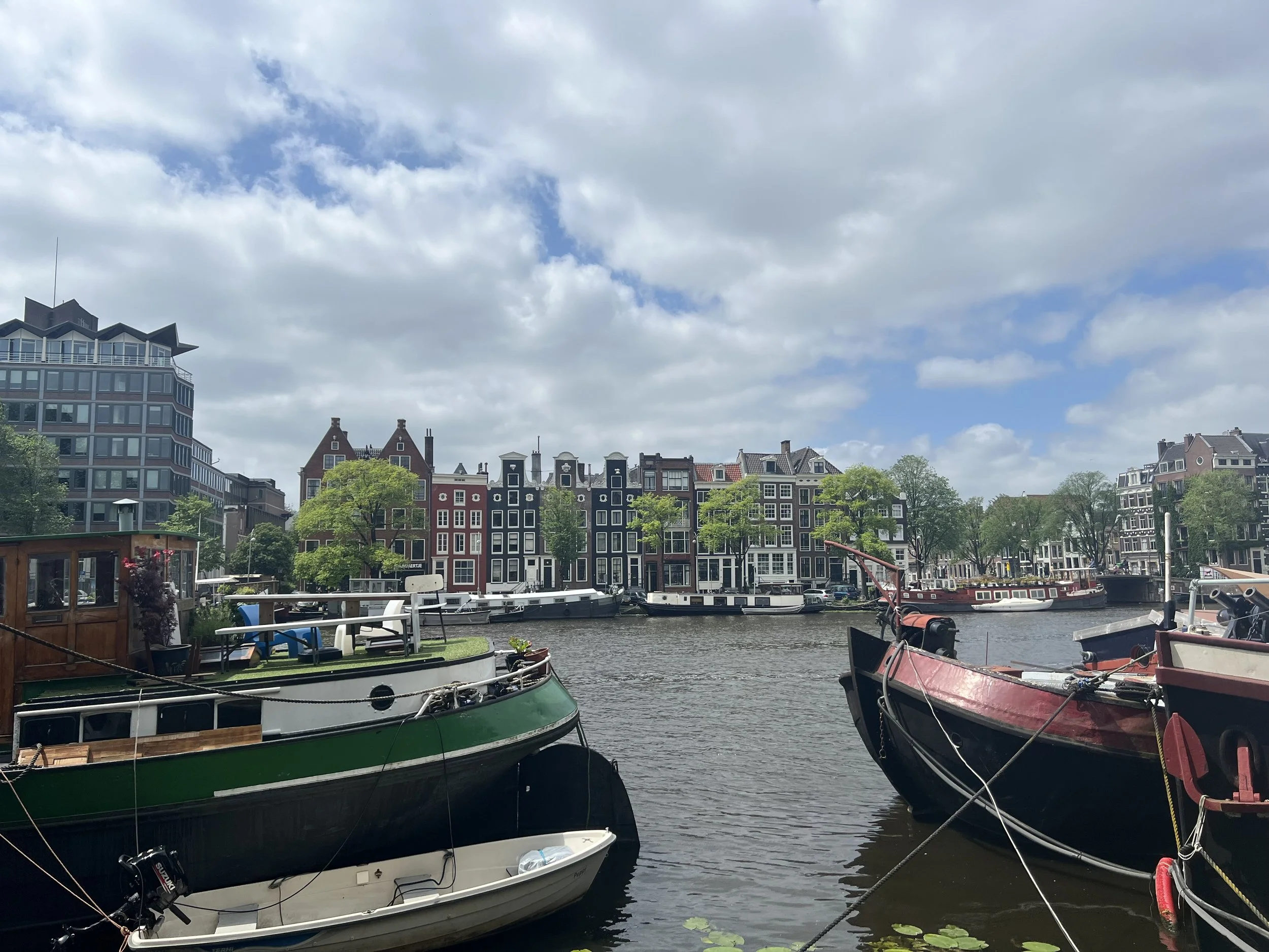 Boats docked along a canal with residential buildings and trees in the background under a partly cloudy sky.