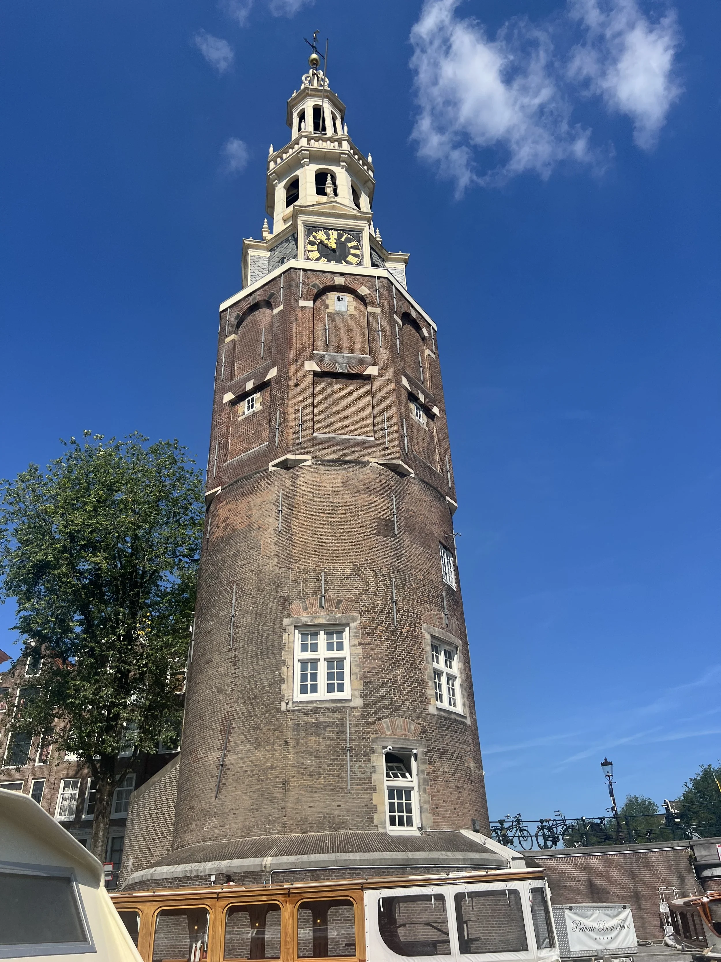 A tall, historic brick tower with white accents, windows, and a clock near the top, under a blue sky with some clouds, with trees and bicycles nearby.