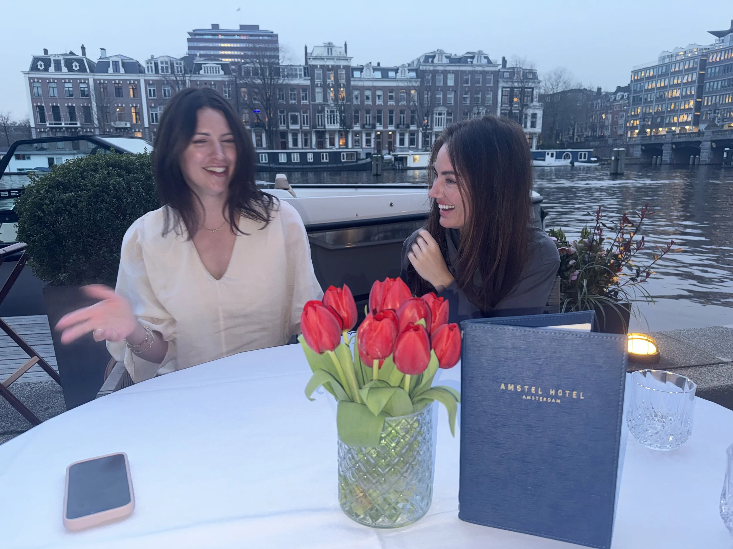 Two women sitting at a table outdoors near a canal, with European-style buildings in the background, in Amsterdam. The table has a vase of red tulips, a smartphone, and a menu.
