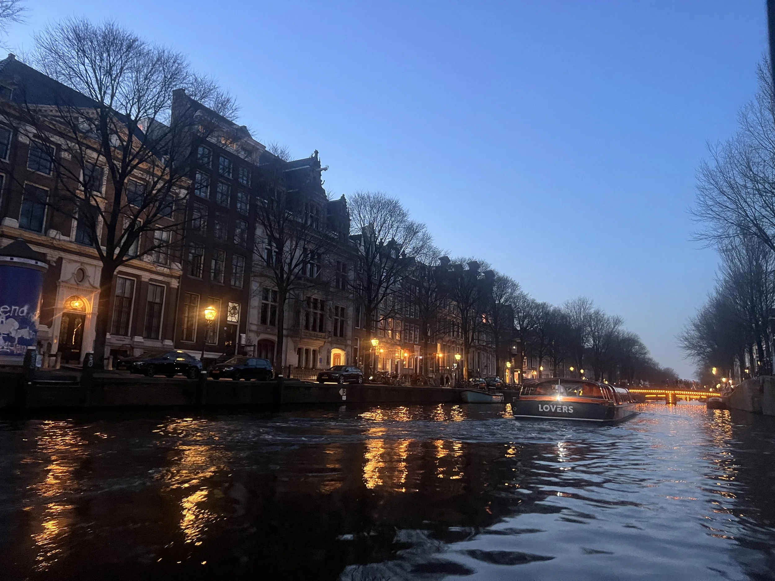 An Amsterdam canal scene during sunset. Illuminated buildings and street lamps at dusk, boat on water with reflected lights, trees without leaves lining the waterway.