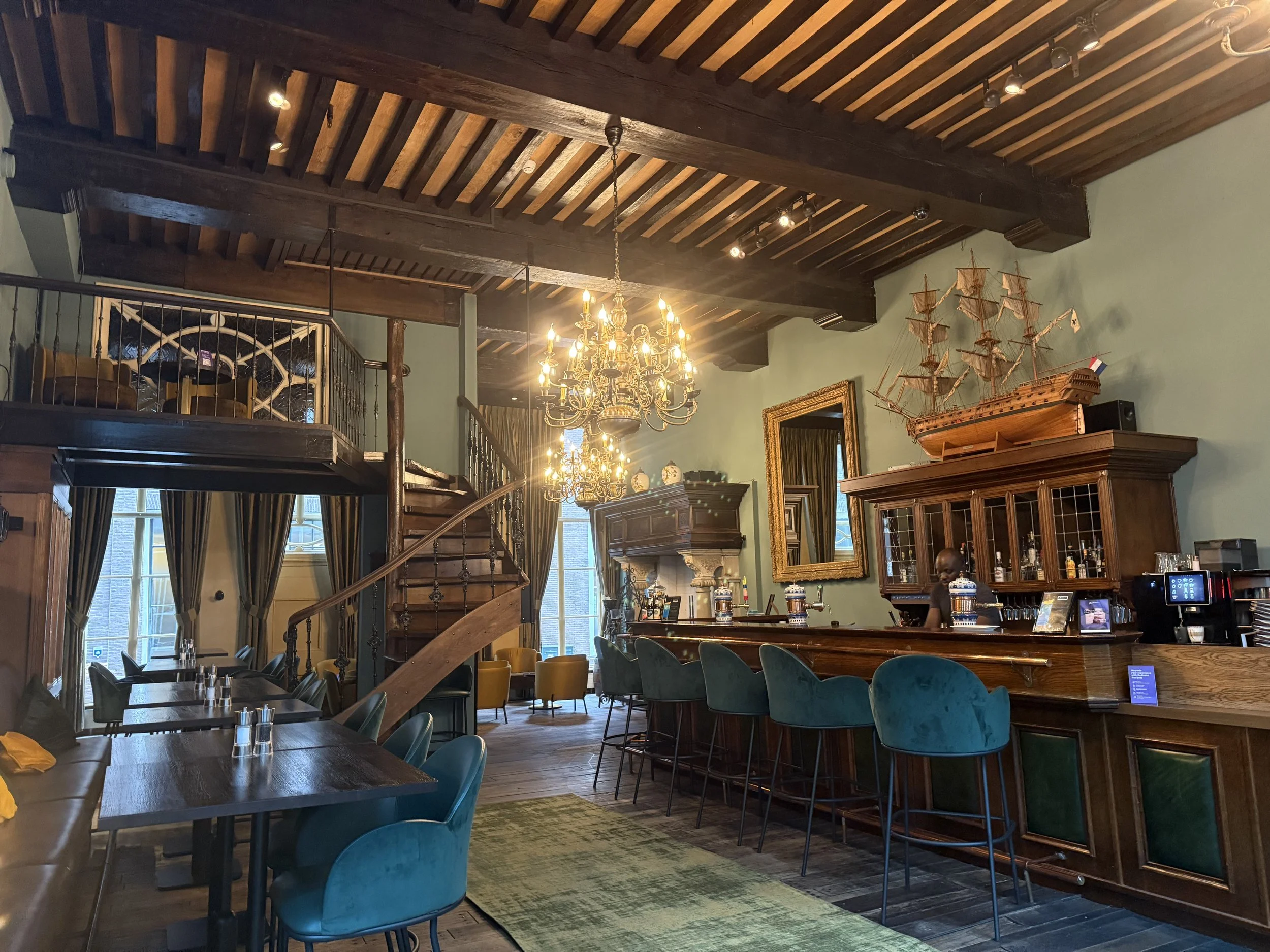 Interior of a restaurant with a high wooden ceiling, a chandelier, and a ship model on display. There are large windows with curtains, a bar area with barstools, and seating arrangements including blue velvet chairs and a long wooden table with chairs.