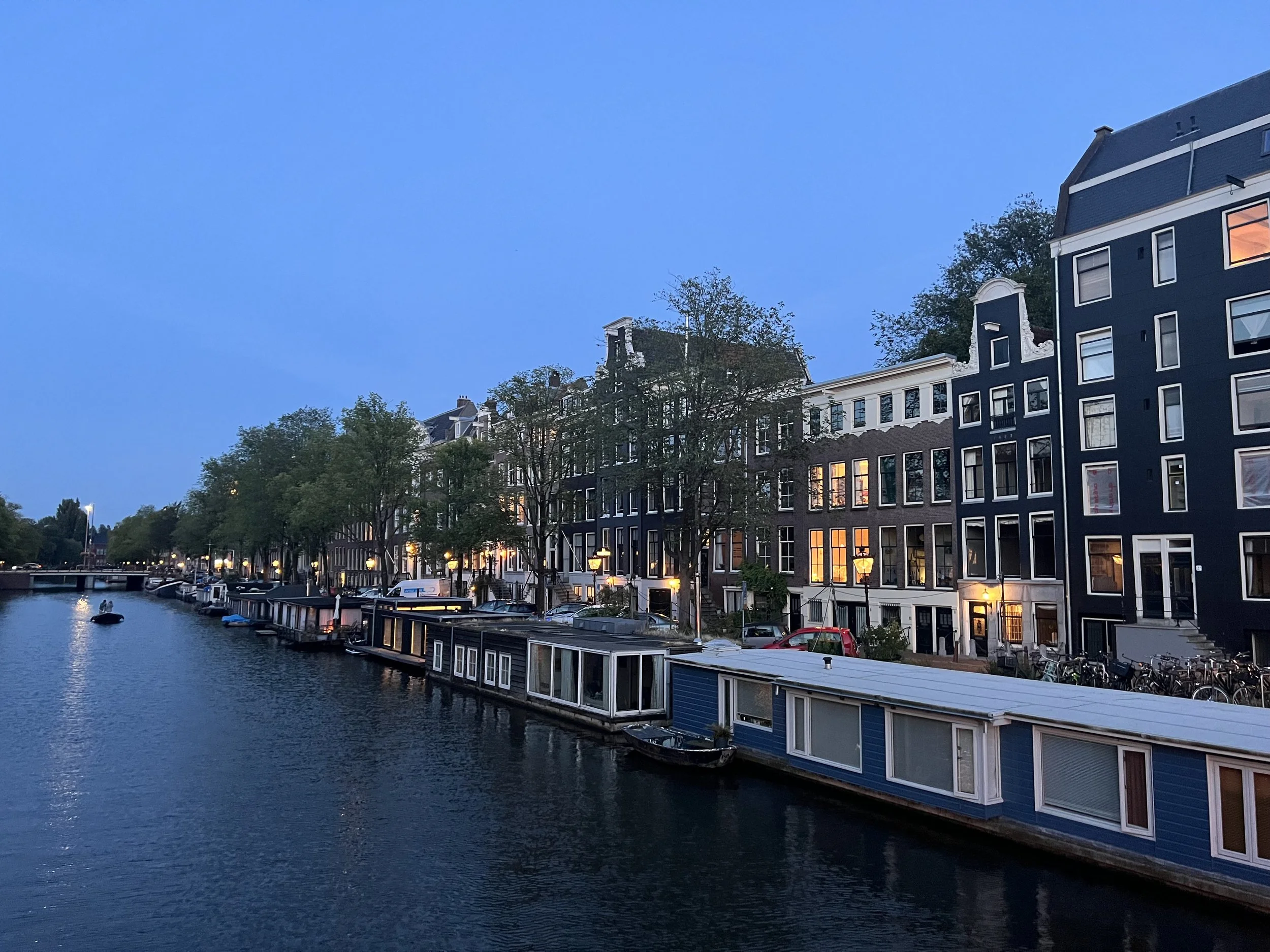 View of a canal with boats, historic buildings, trees, and street lamps in a European city during early evening or twilight.