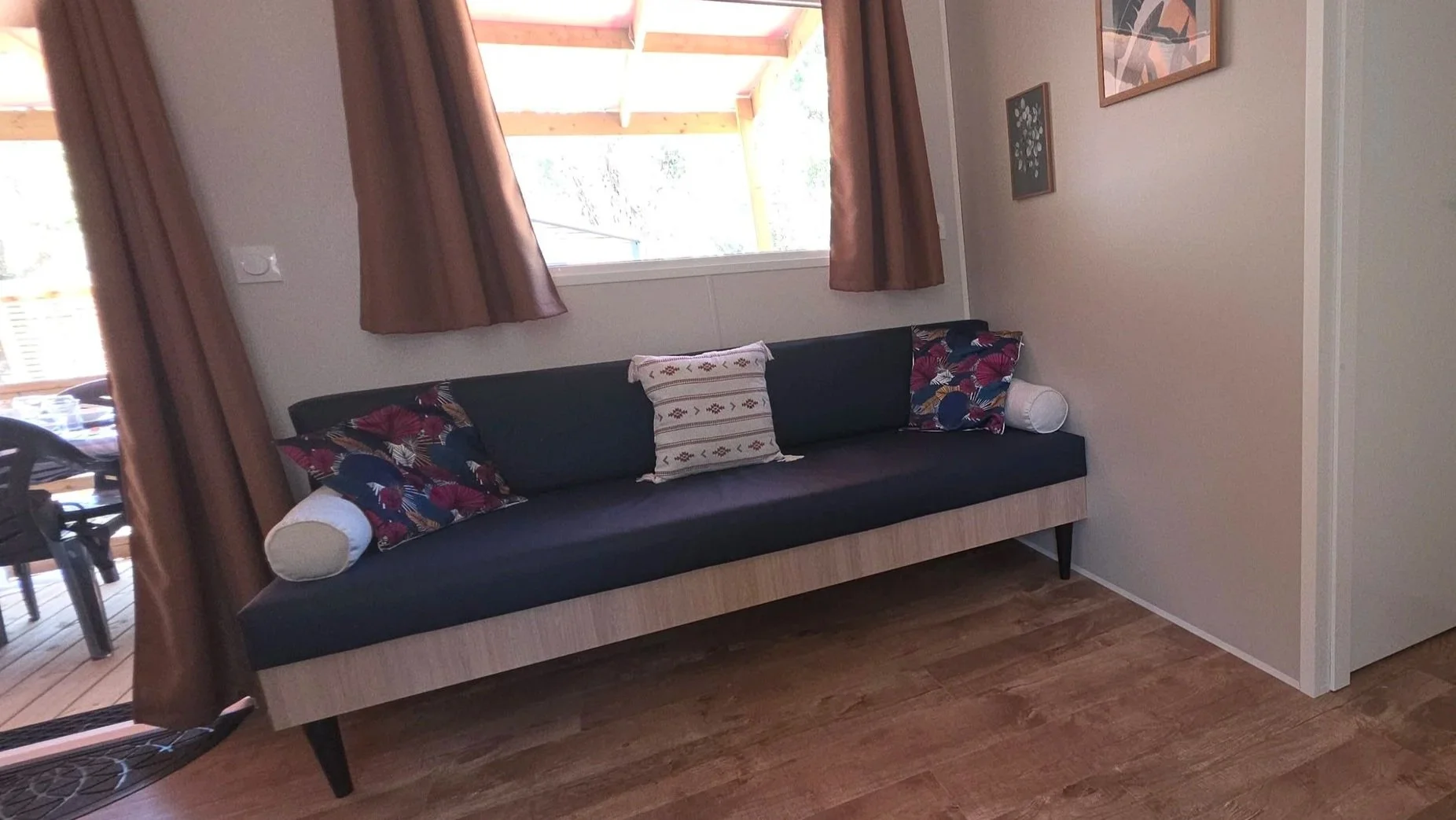 Intérieur d'une pièce avec un canapé noir, décorée avec des coussins colorés et encadrés sur le mur, fenêtres avec rideaux marrons et parquet en bois.