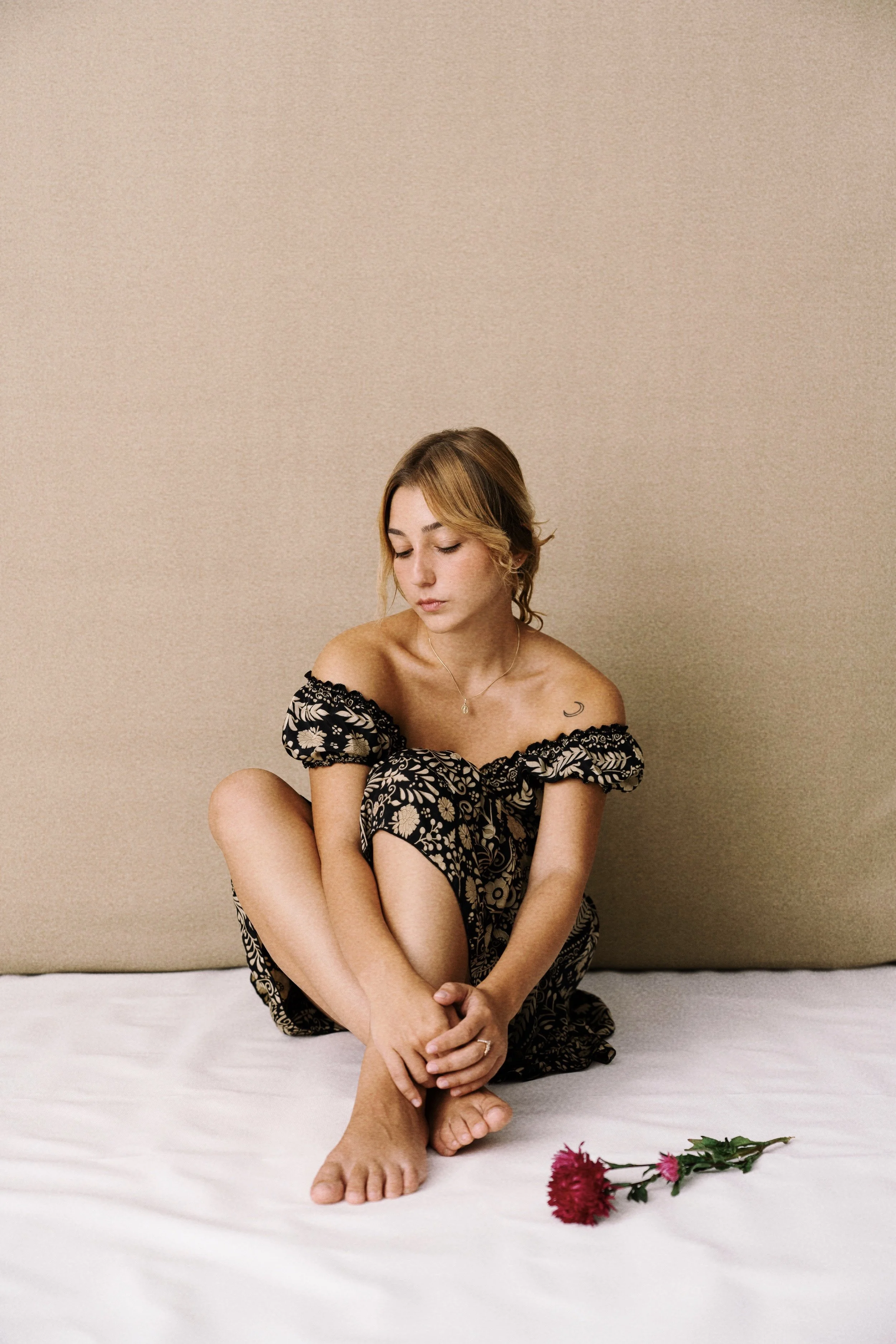 A woman sitting on a white surface against a beige wall, wearing a black and beige floral dress, with rose flowers and a cup nearby.