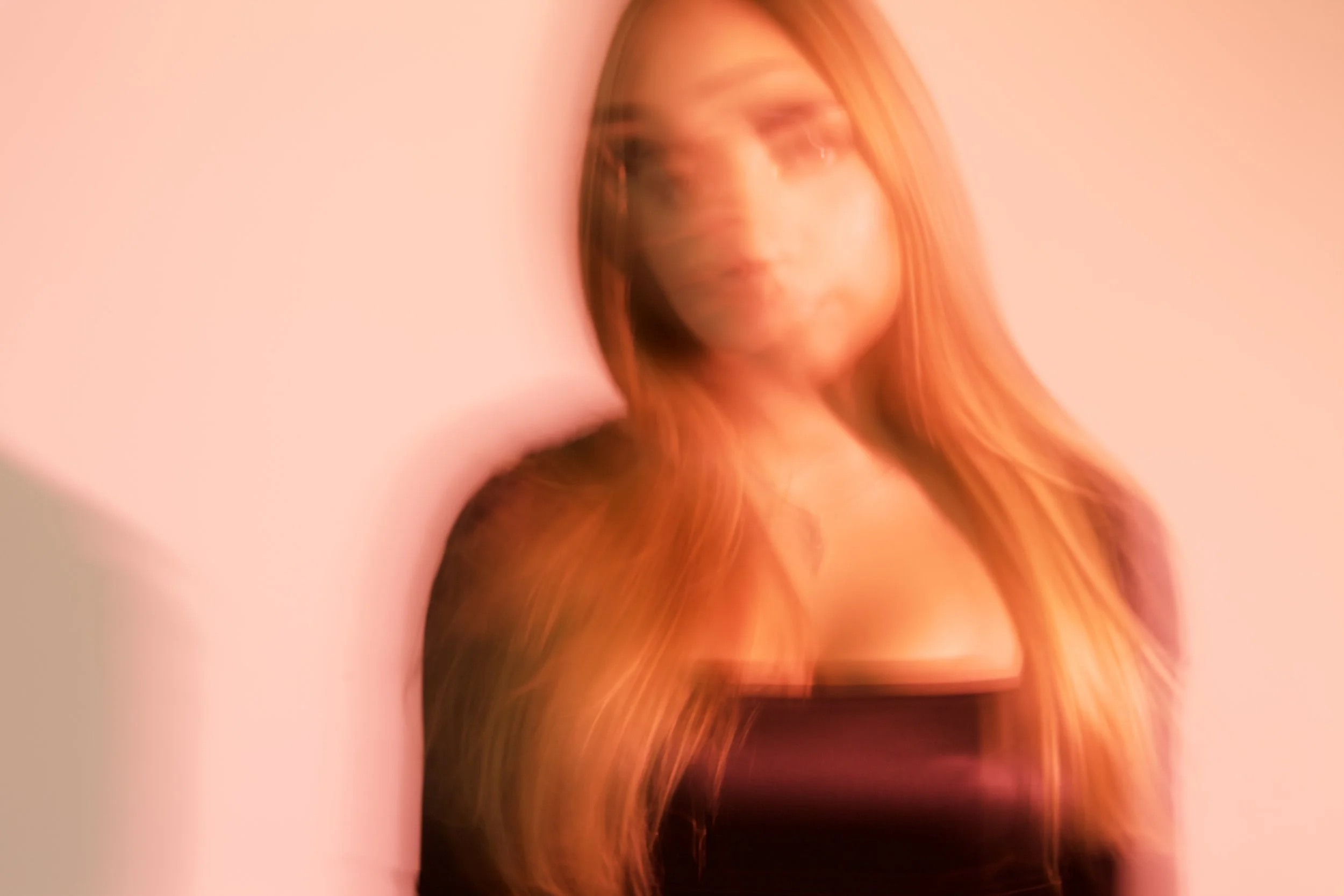 A woman with long hair and makeup, leaning against a pink background, with a blurred, artistic effect.