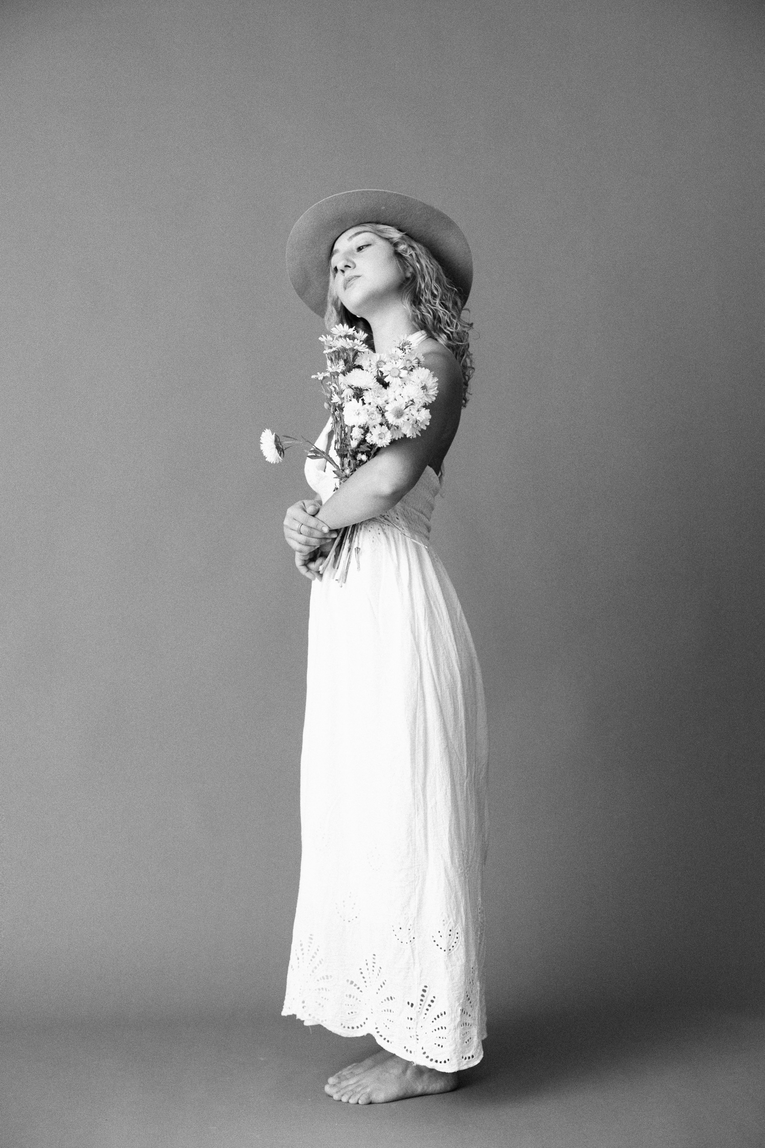 A woman standing barefoot on a plain background, holding a bouquet of flowers, wearing a wide-brimmed hat and a long skirt with embroidery details.