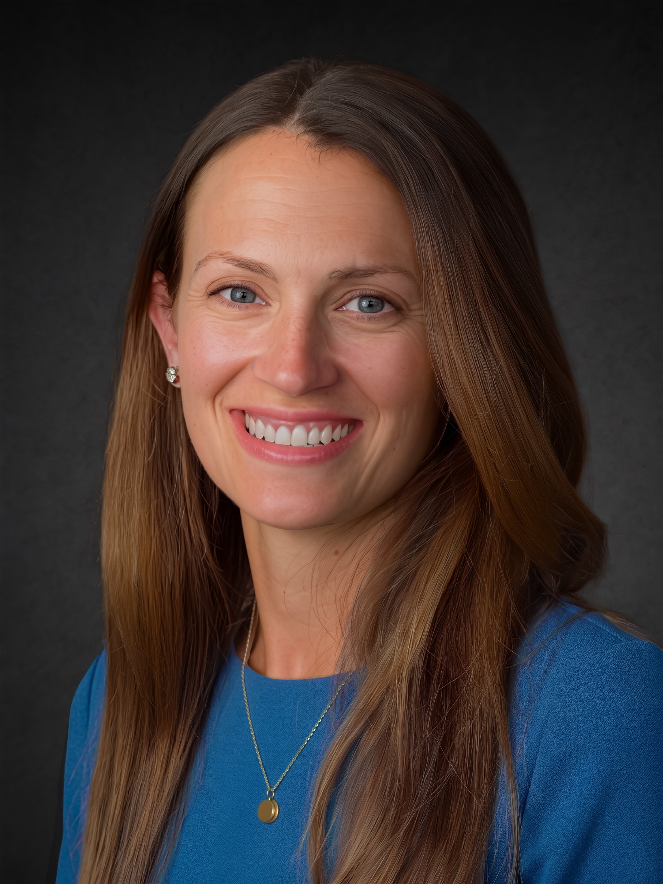 Close-up portrait of smiling woman with long light brown hair, blue eyes, wearing a blue top, gold necklace with a round pendant, and a small earring, against a dark background.
