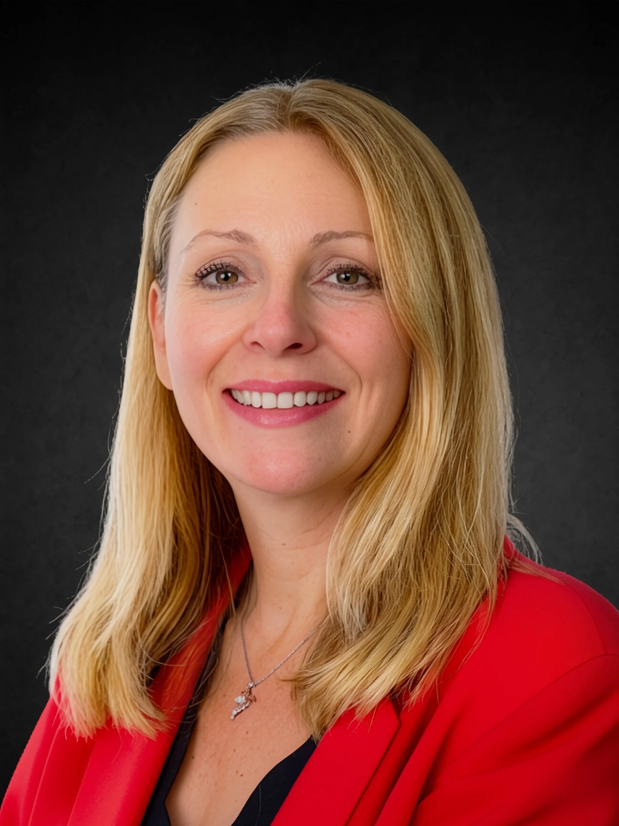Close-up photo of a smiling woman with blonde hair, wearing a red blazer, black top, and a necklace, against a black background.