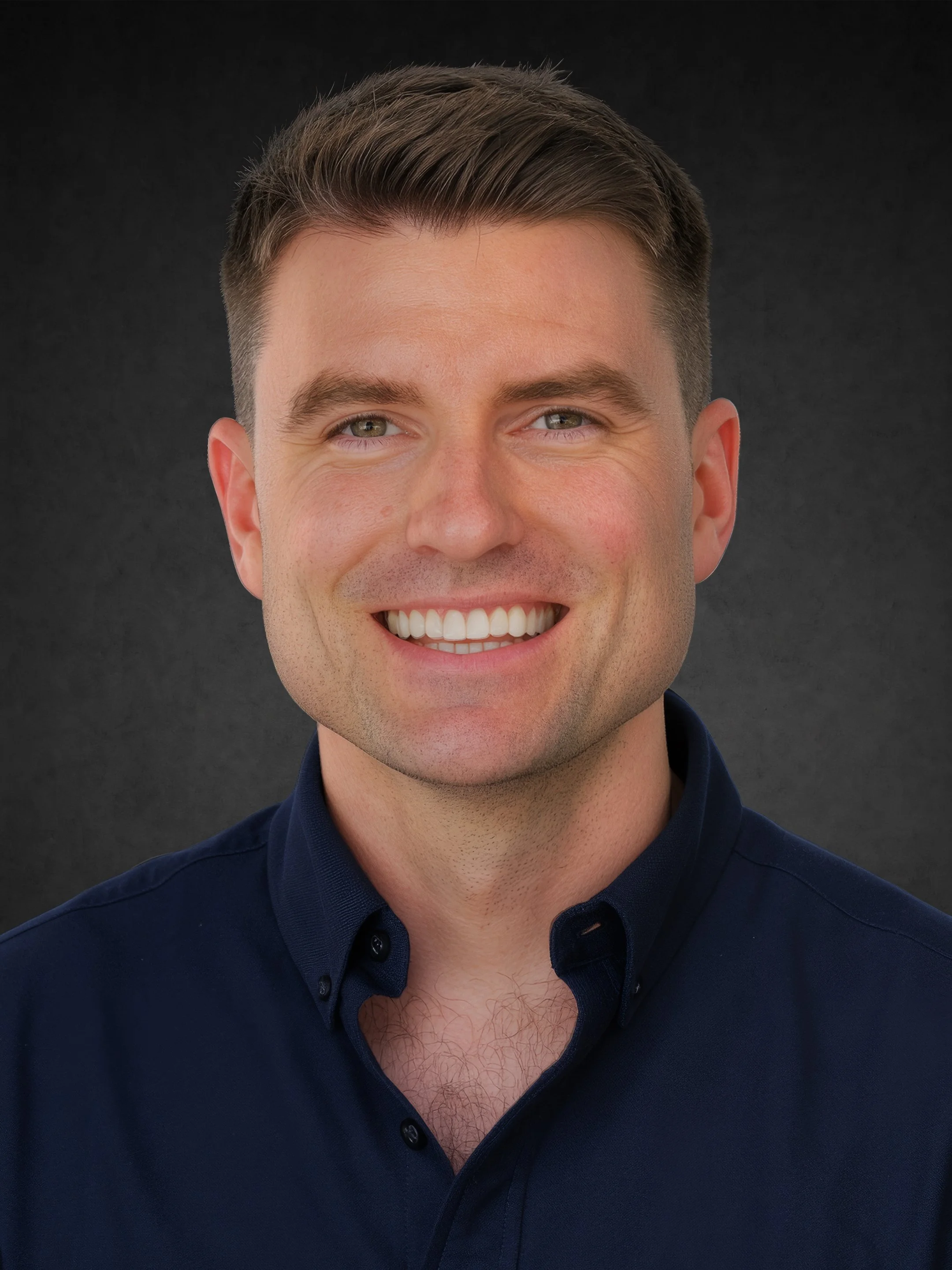 Close-up of a smiling man with short brown hair, light skin, wearing a dark blue collared shirt against a dark background.