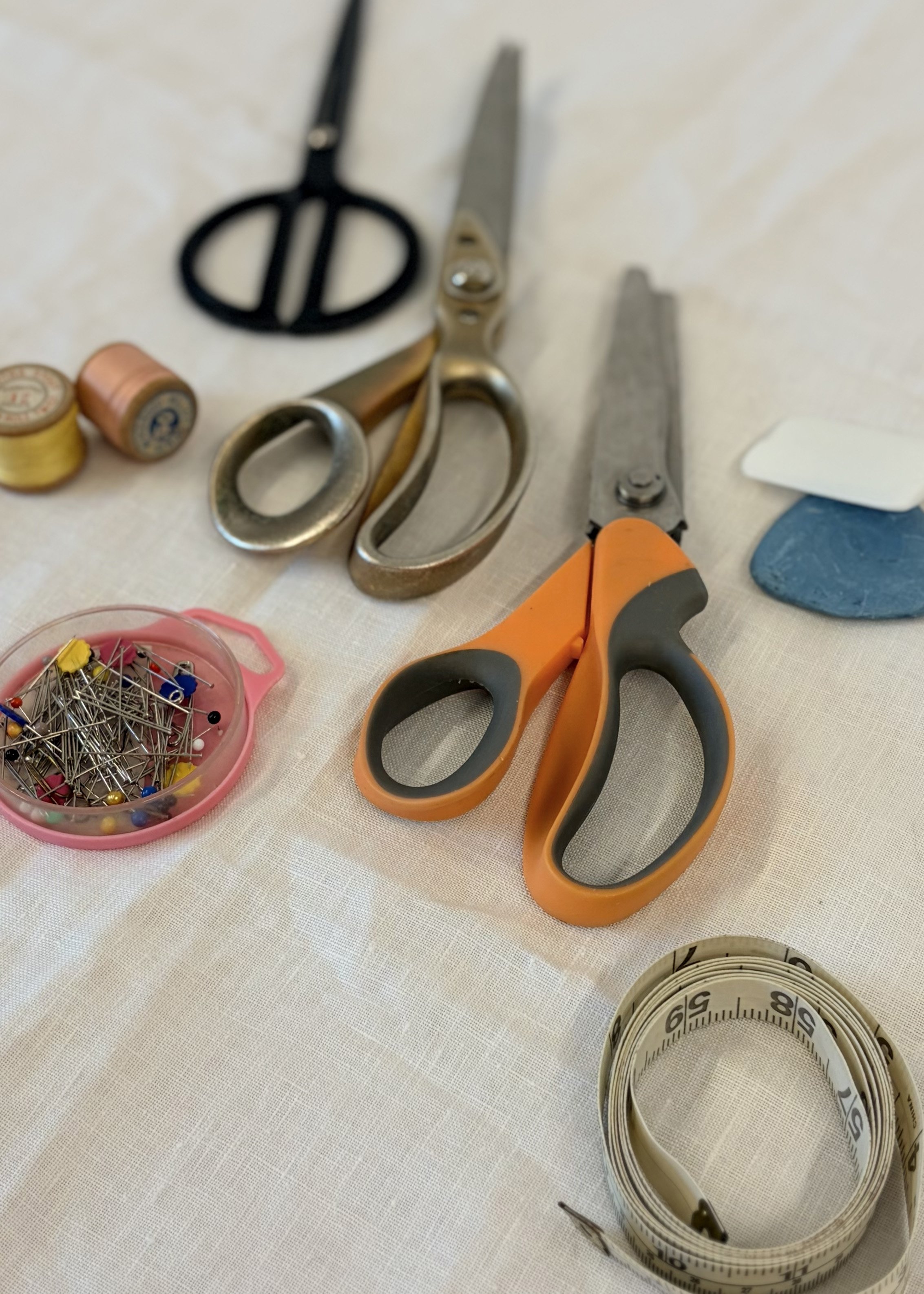 Sewing tools including a pair of scissors, a measuring tape, a small container of pins, and spools of thread on a white fabric surface.