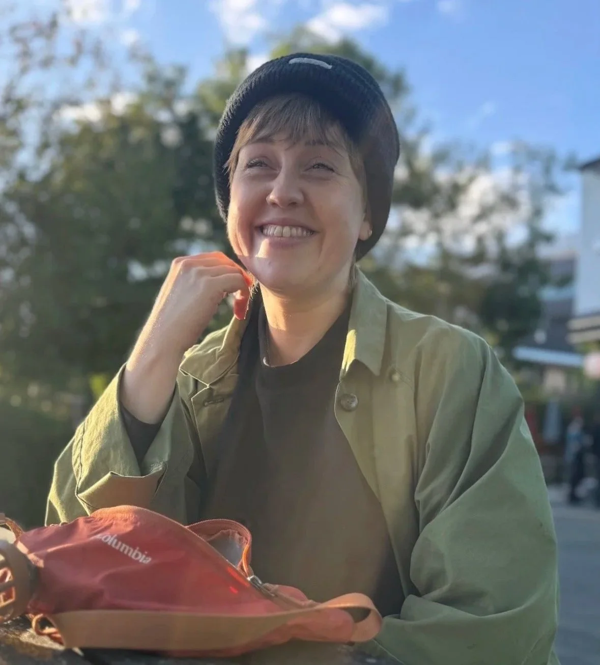 A woman with short hair wearing a black beanie, green jacket, and black shirt sitting outdoors, smiling and looking upward, with a red Columbia backpack in front of her, in a park or outdoor area with trees and buildings in the background.