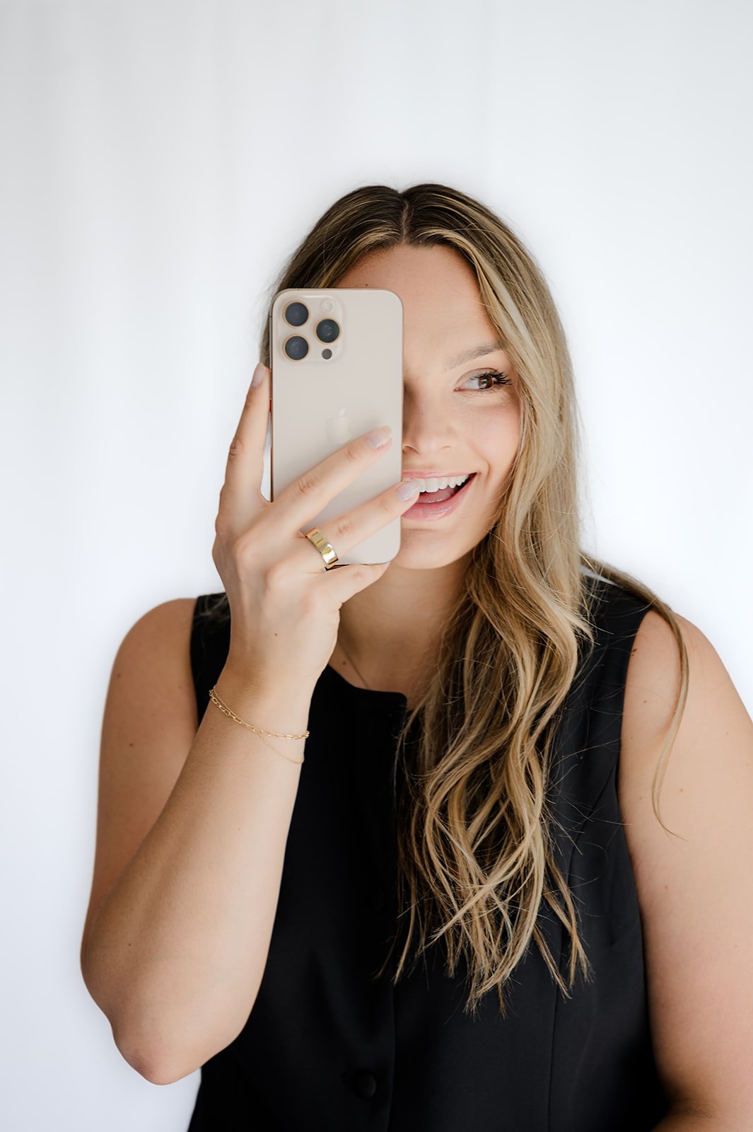 A woman with long, wavy blonde hair takes a selfie with a smartphone, smiling and looking to the side, against a plain white background. She wears a black sleeveless top and gold jewelry.