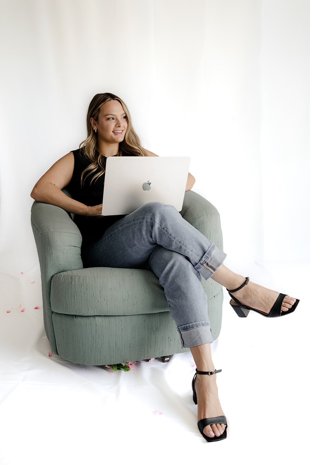 A woman sitting on a green armchair, working on a silver MacBook, with crossed legs, in a bright room with a white background.