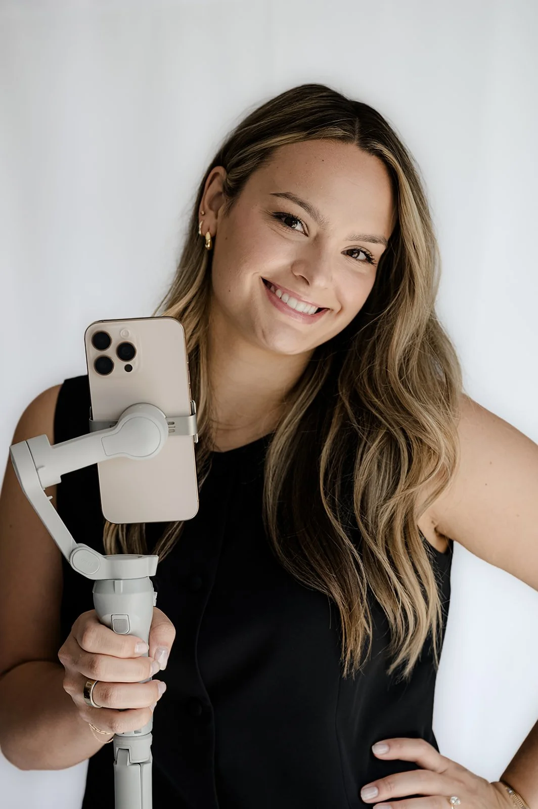 A young woman with long, wavy hair takes a selfie using a smartphone mounted on a selfie stick. She is smiling and wearing a black sleeveless top, with earrings and rings.