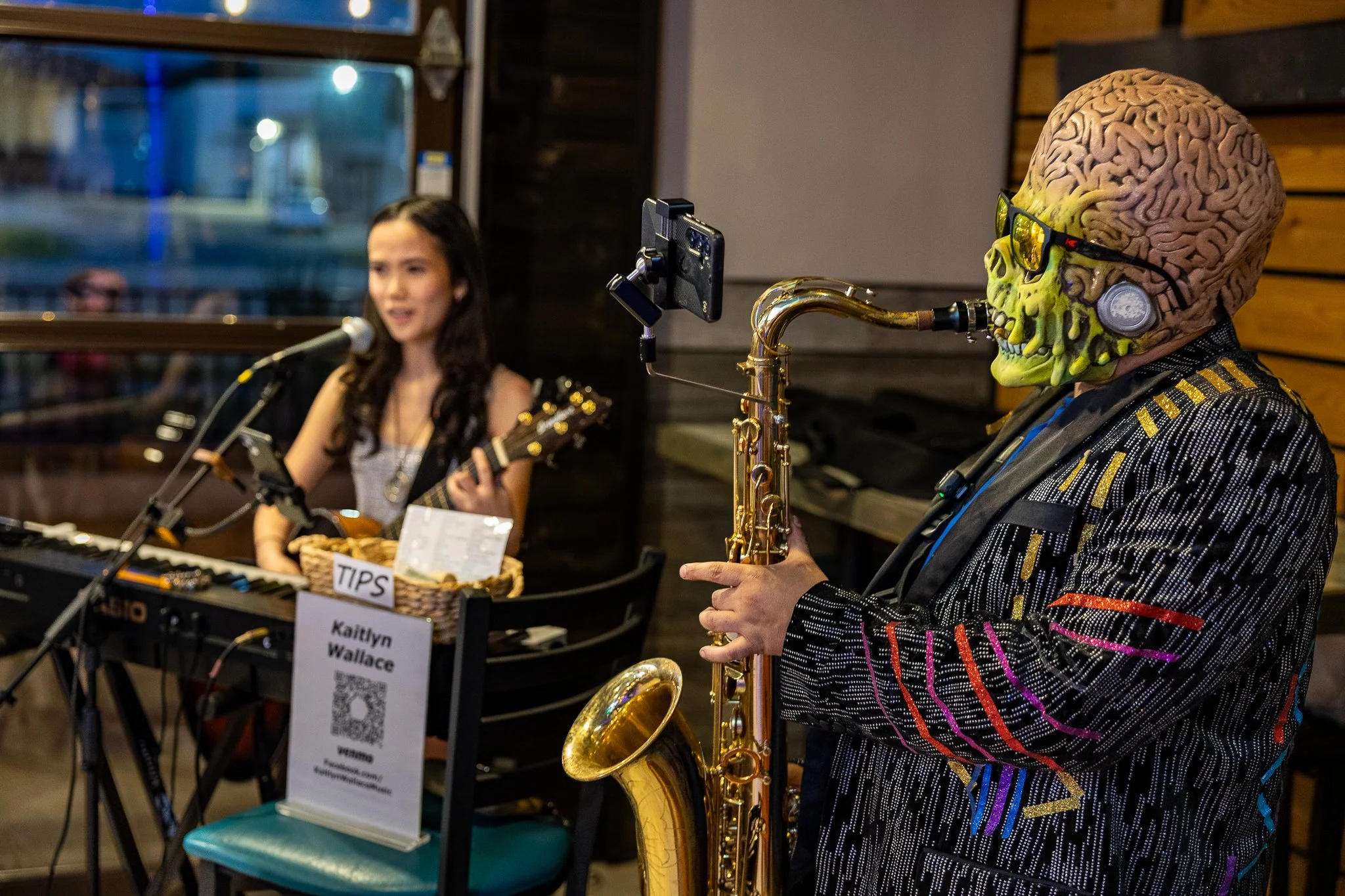 A woman playing keyboard and singing into a microphone and a skeleton wearing sunglasses playing a saxophone during a live music performance in a cozy indoor setting.