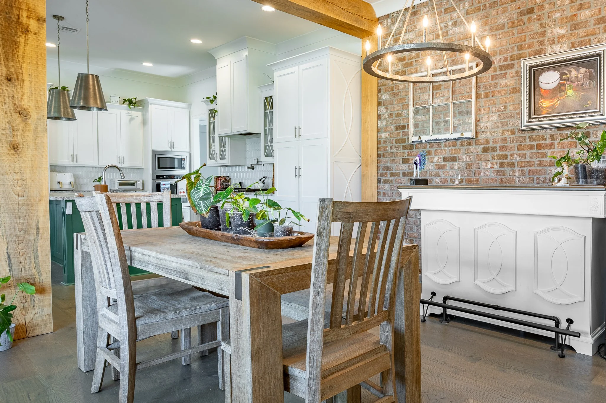 A dining area with a wooden table and chairs, a brick wall with a framed picture of beer glasses, a chandelier, and a kitchen in the background with white cabinets and appliances.