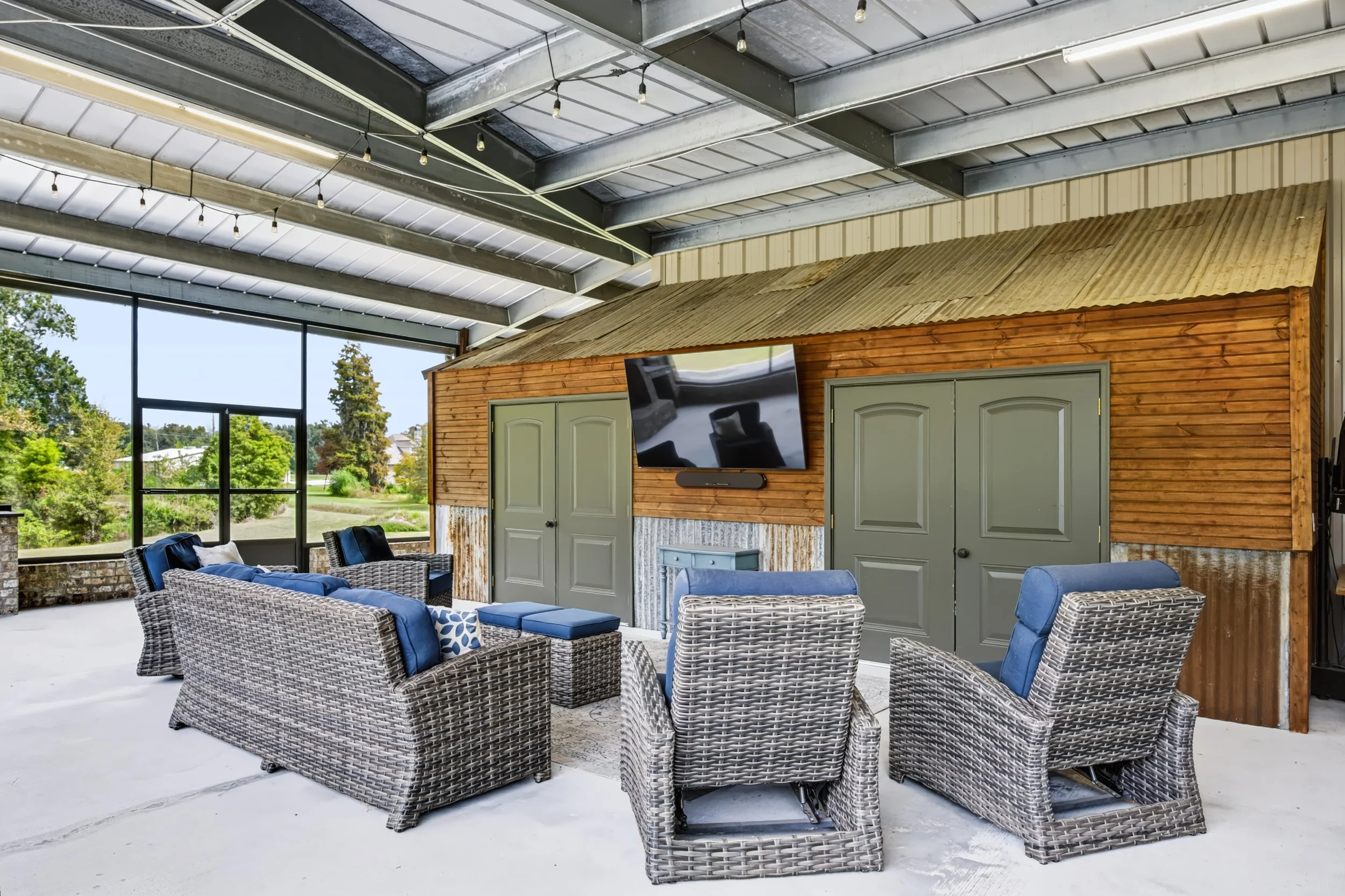 An outdoor patio with wicker furniture including chairs and a sofa, a wooden shed with closed doors, a flat-screen television mounted on the shed wall, string lights hanging from the metal roof, and a view of trees and a grassy landscape outside.