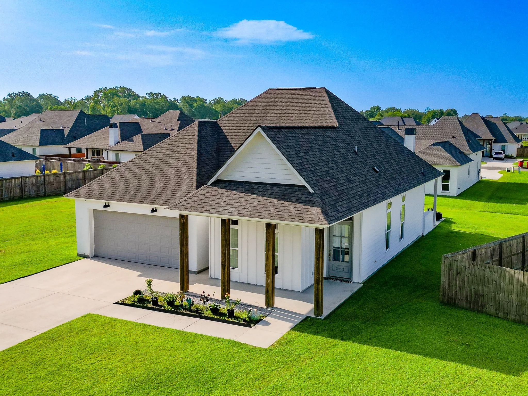 A suburban backyard with a white house, a concrete driveway, a fenced yard, and neighboring houses with brown roofs under a bright blue sky.