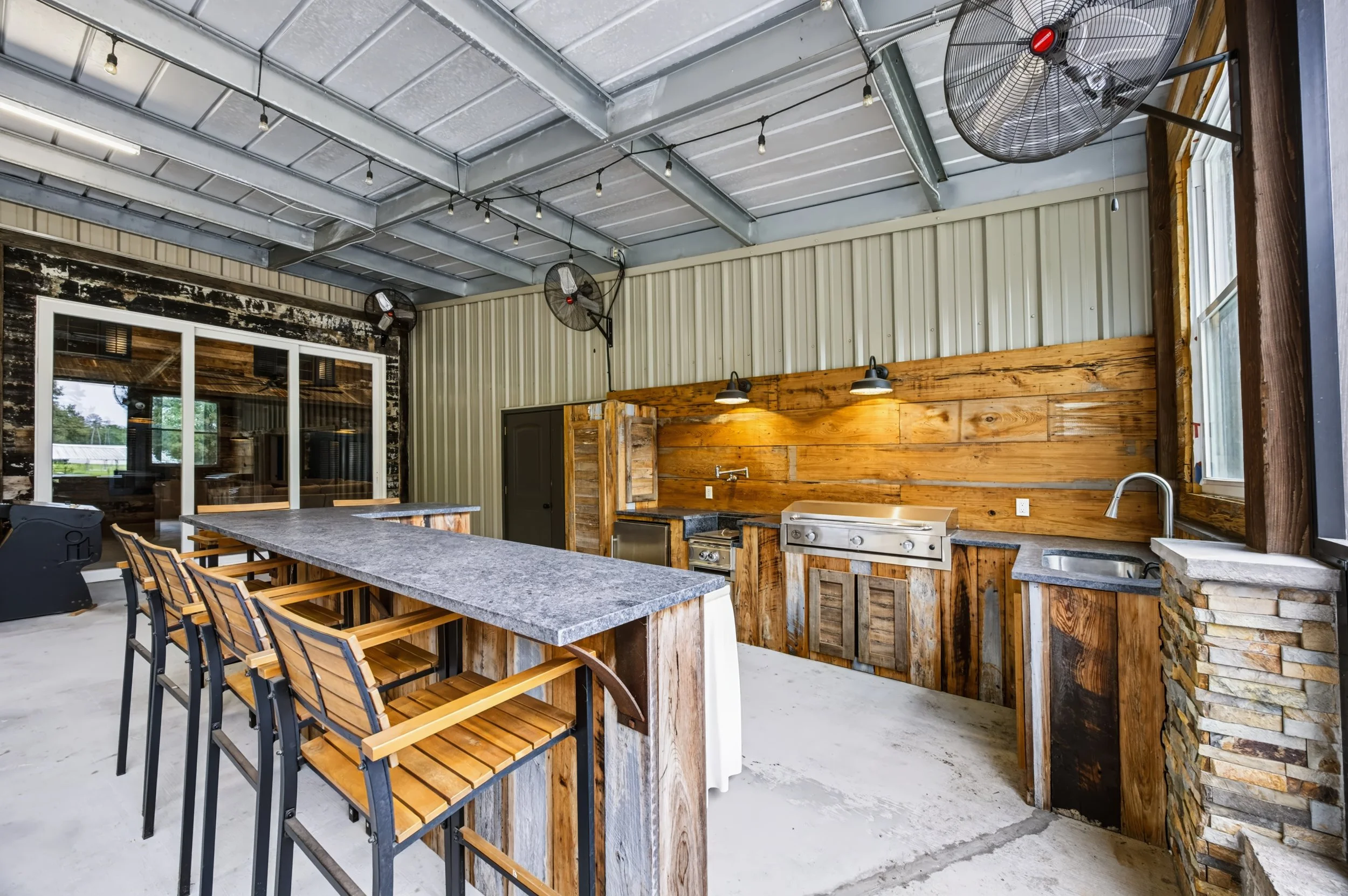 Patio area with a large concrete table and wooden chairs, enclosed by metal and wooden walls, equipped with ceiling fans, outdoor lighting, and a stainless steel grill.