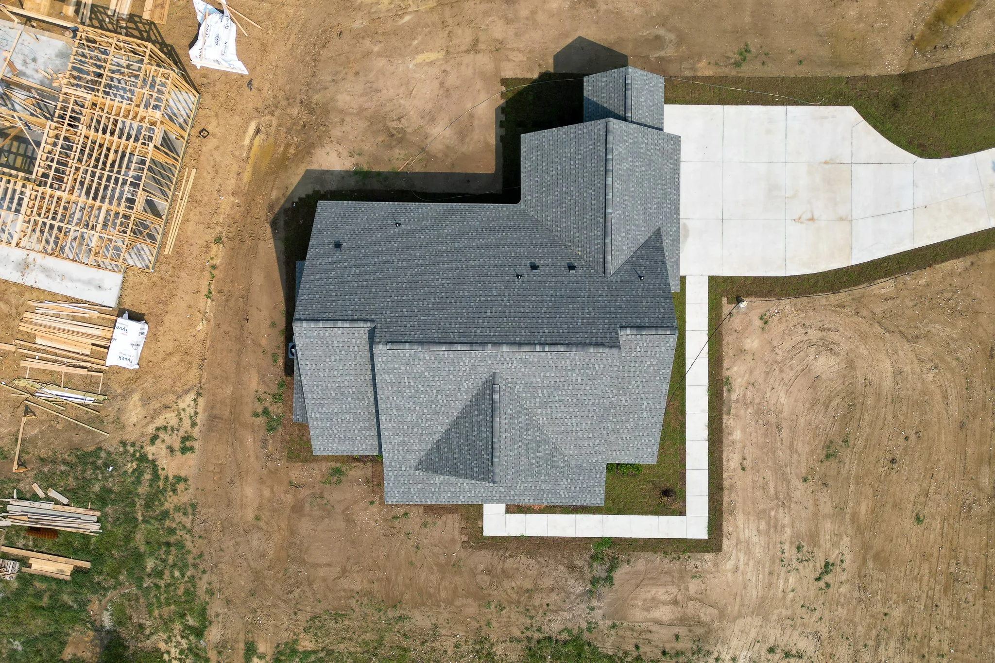 An aerial view of a house under construction with a partially built wooden framework on the left, a finished building with a dark gray shingle roof in the center, and a freshly poured concrete driveway and walkway on the right. The surrounding area i