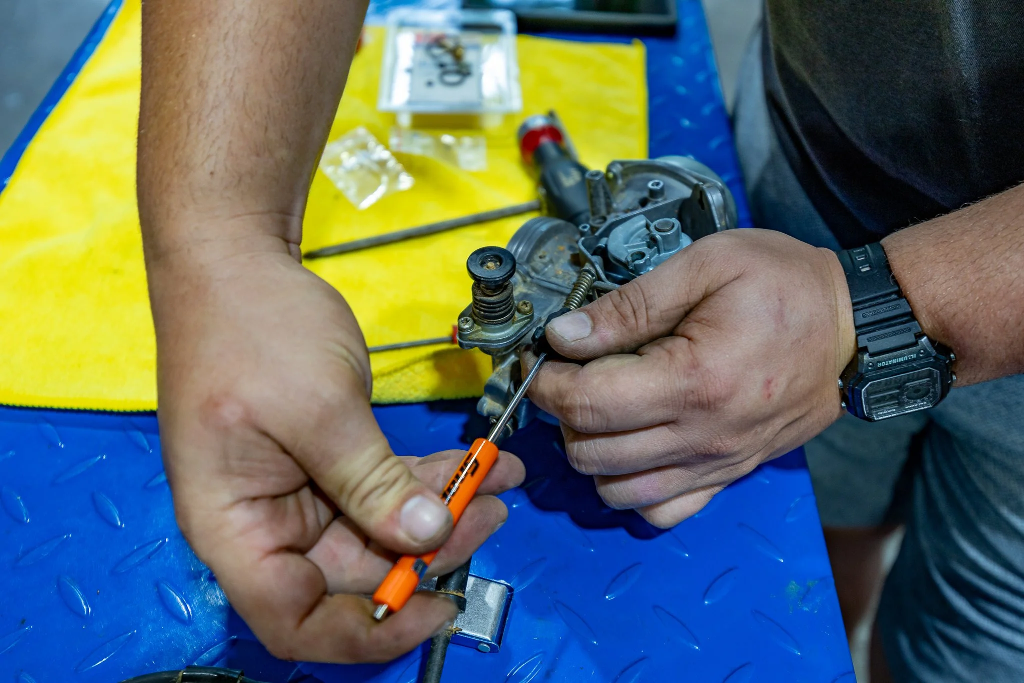 A person working on a mechanical component, possibly a carburetor, with small tools and parts on a yellow and blue workspace.