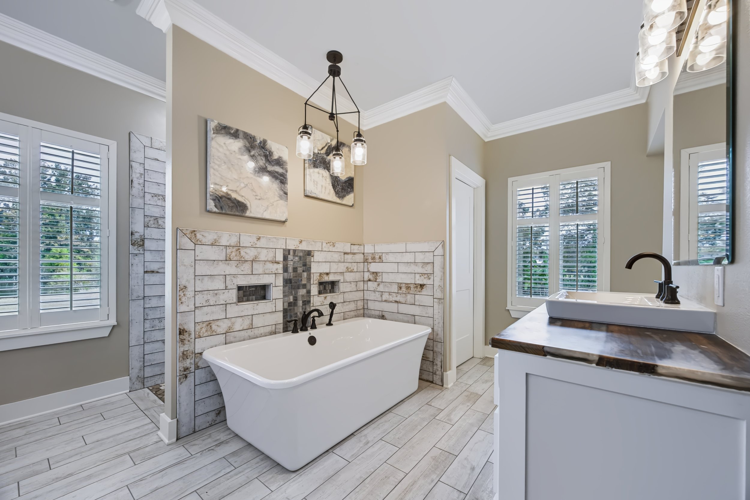 A bright bathroom with a freestanding bathtub with black fixtures against a wall of beige tiles featuring a decorative pattern. There are two windows with white shutters, a white vanity with a sink and black faucet, and two modern light fixtures hang