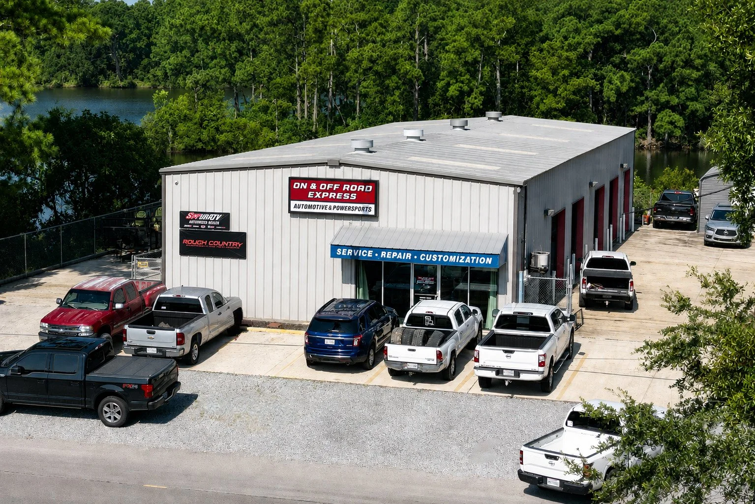 An automotive repair and customization shop with several trucks and cars parked in front of the building. Signs indicate services including service, repair, customization, and motorsports.